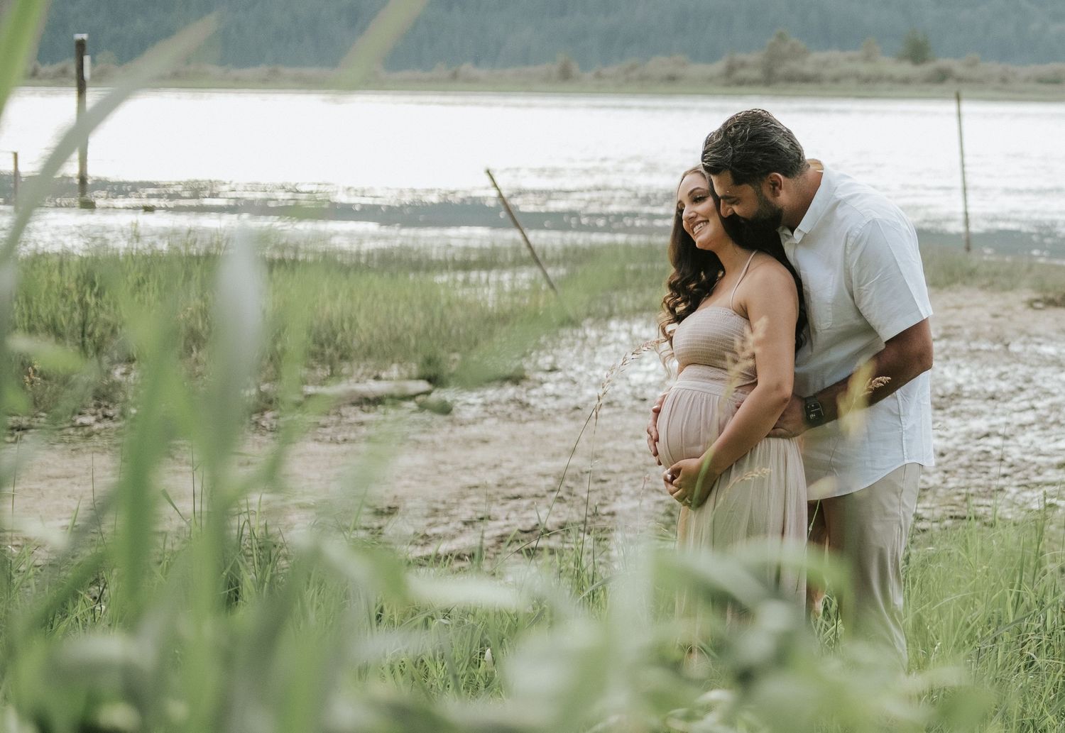 Pregnant couple embracing near a serene lake, surrounded by lush green grass during a maternity photo session.
