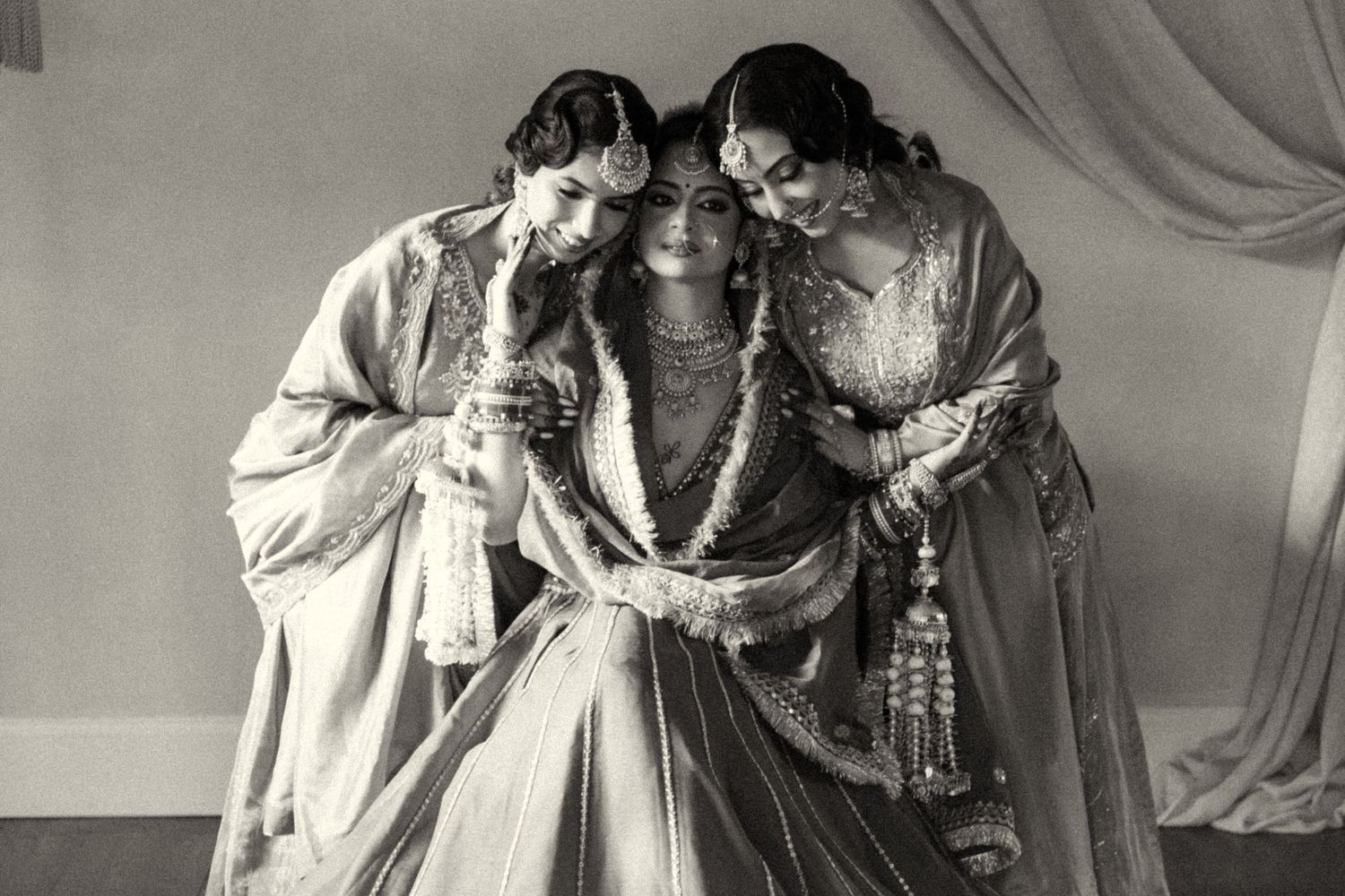 Black and white photo of Indian bride flanked by two women in elegant ethnic attire with floral garlands.
