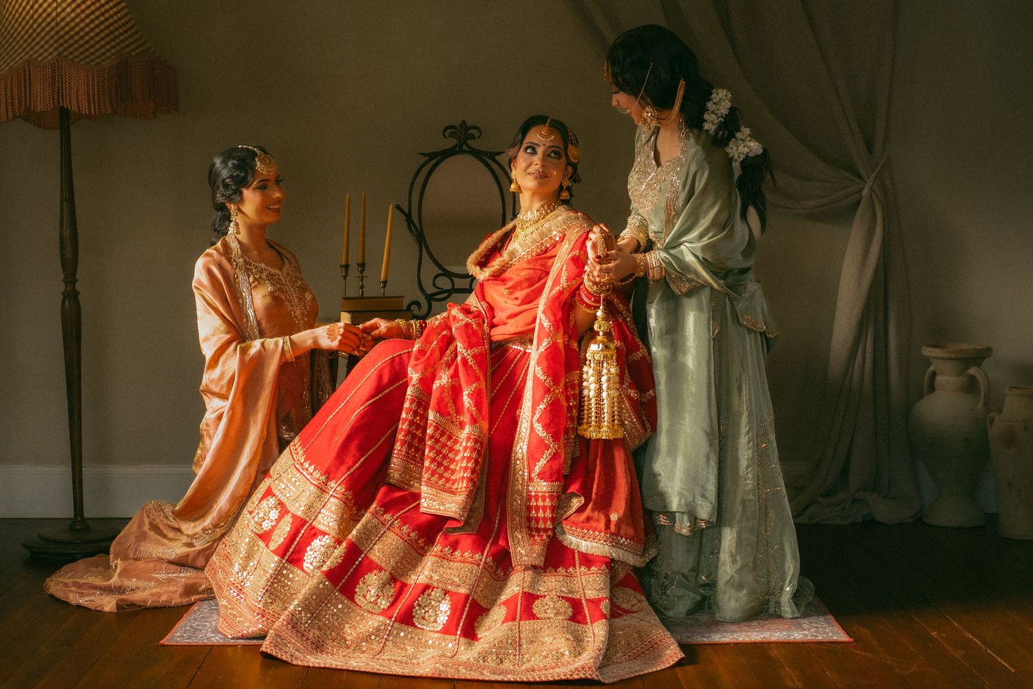 Indian bride in red and gold lehenga assisted by bridesmaids in soft moody studio setting.