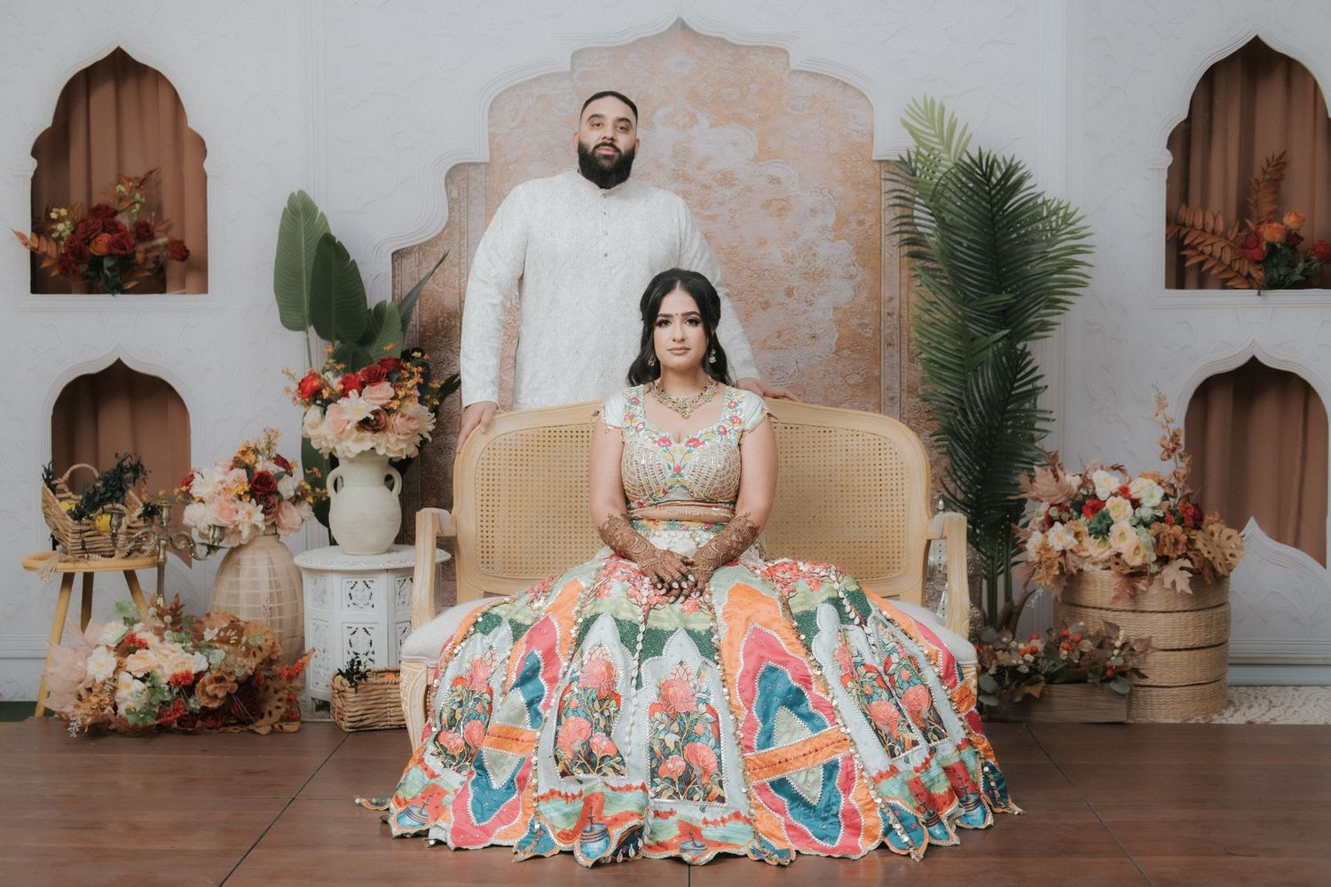 Indian wedding couple posing, bride seated in vibrant floral lehenga, groom standing in white sherwani behind her.