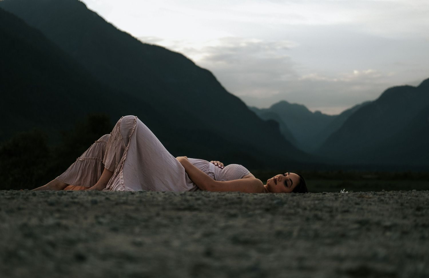 A woman in a flowing white dress reclines on an open field with dramatic mountain silhouettes at twilight.