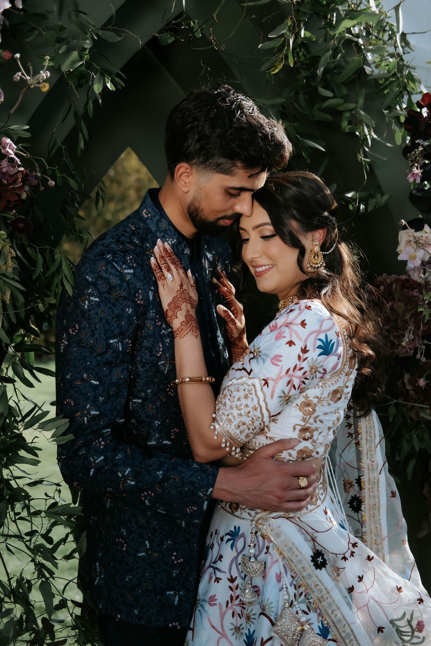 Indian couple embracing tenderly under a dark floral arch at a romantic wedding ceremony.