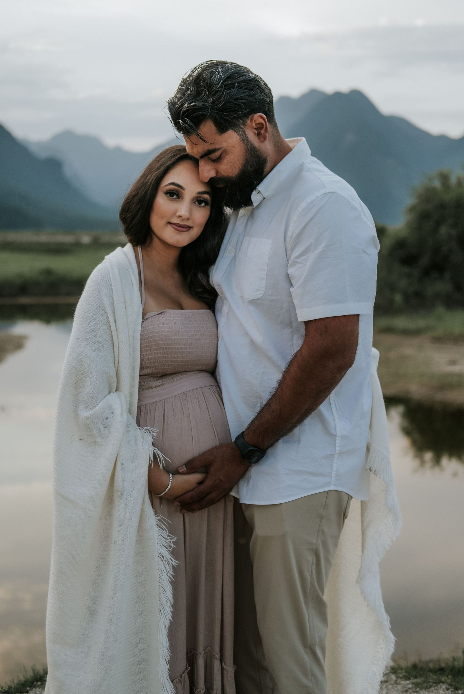 Couple tenderly embracing with man nuzzling pregnant woman's forehead near a serene mountain lake at dusk.