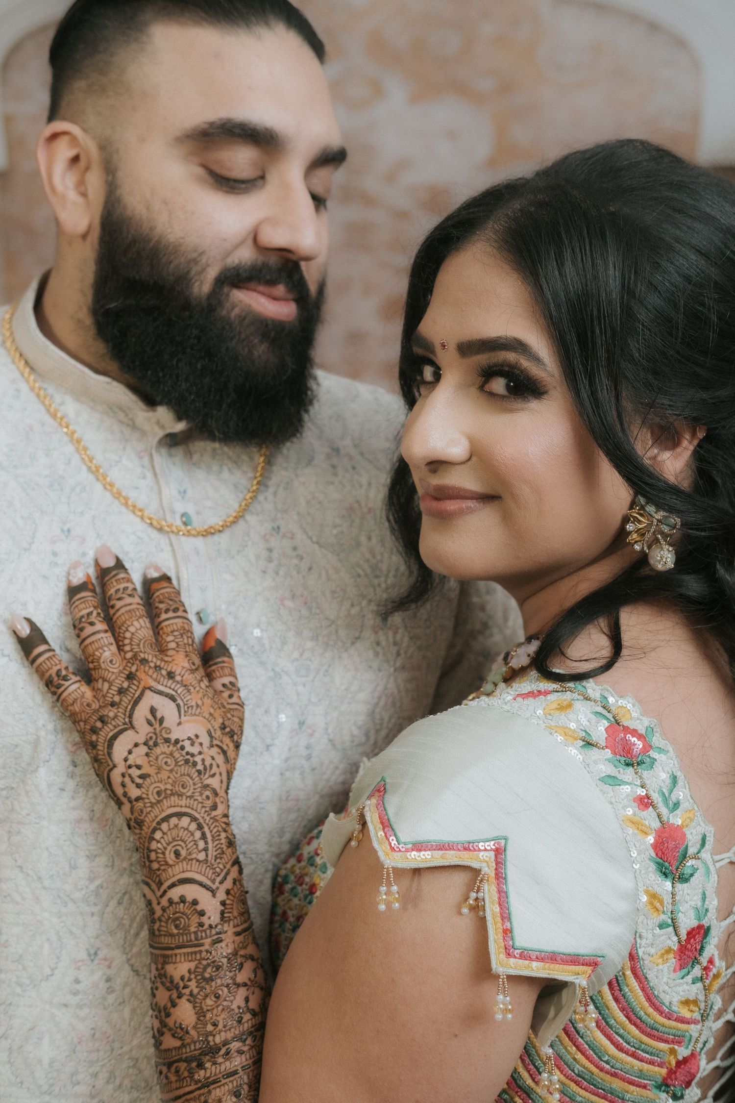 Close-up of Indian bride with intricate mehndi leaning against groom in white sherwani with gold chain.