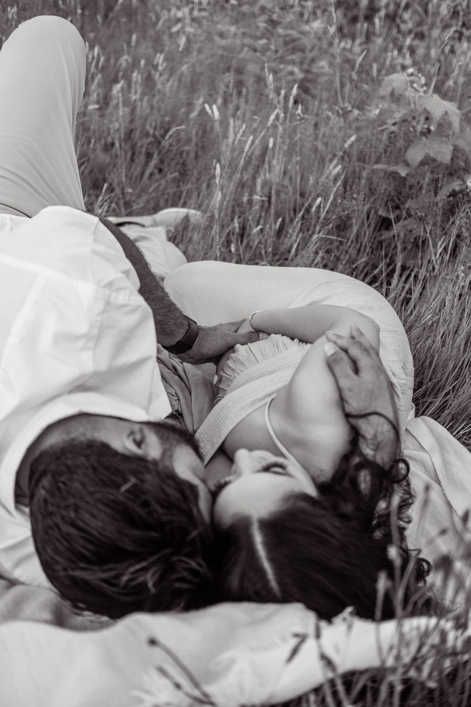 Black and white photo of a couple lying in tall grass, sharing an intimate moment during maternity shoot.