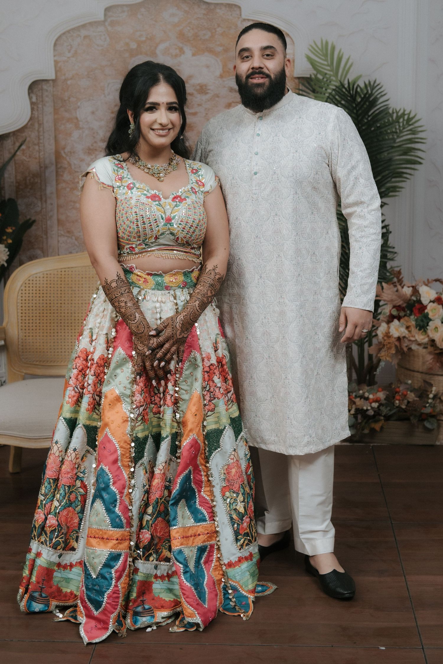 Couple in Indian wedding attire, bride in colorful floral lehenga with mehndi, groom in white kurta pajama.
