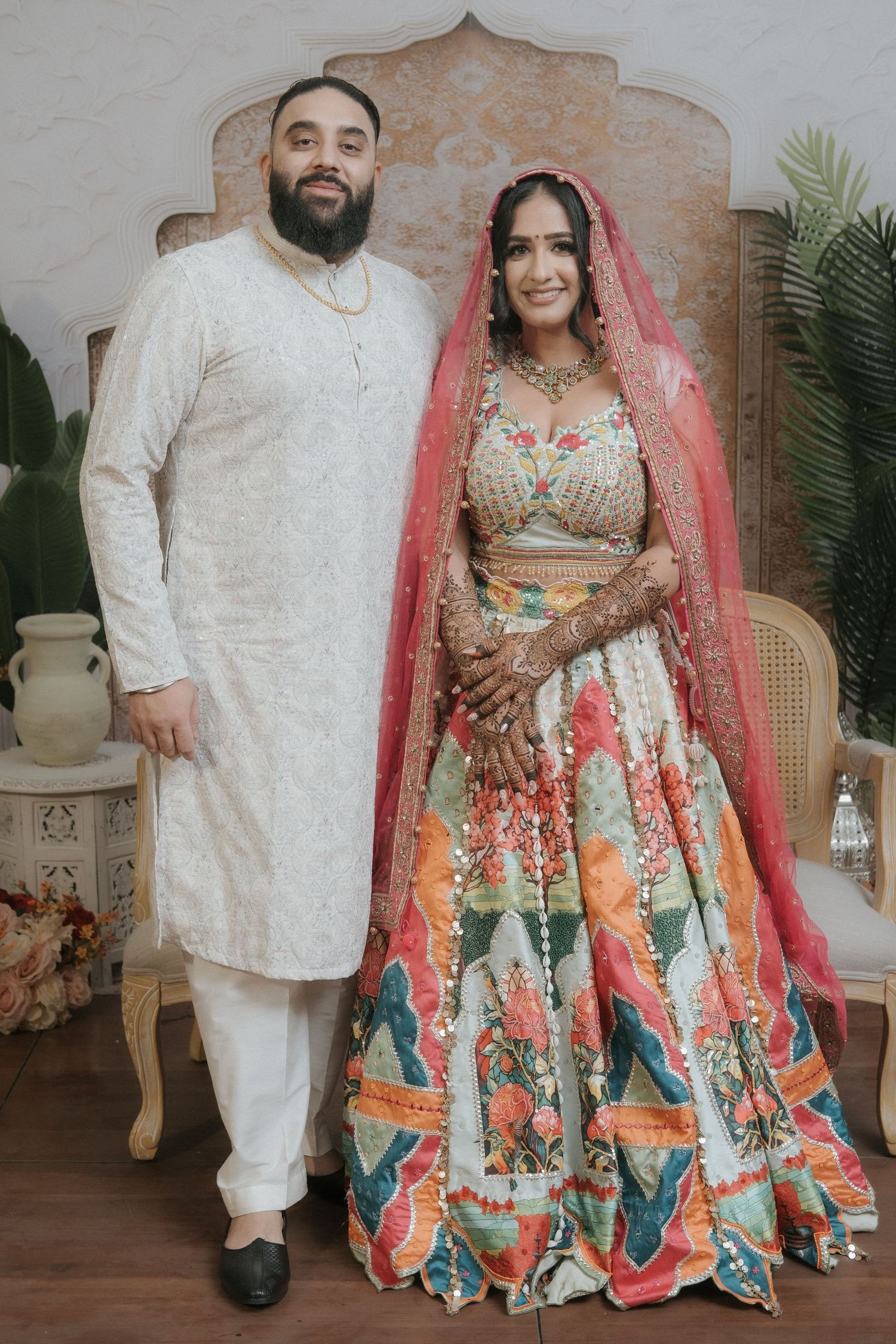 Indian bride in colorful lehenga with pink dupatta and groom in white kurta posing together at wedding ceremony.