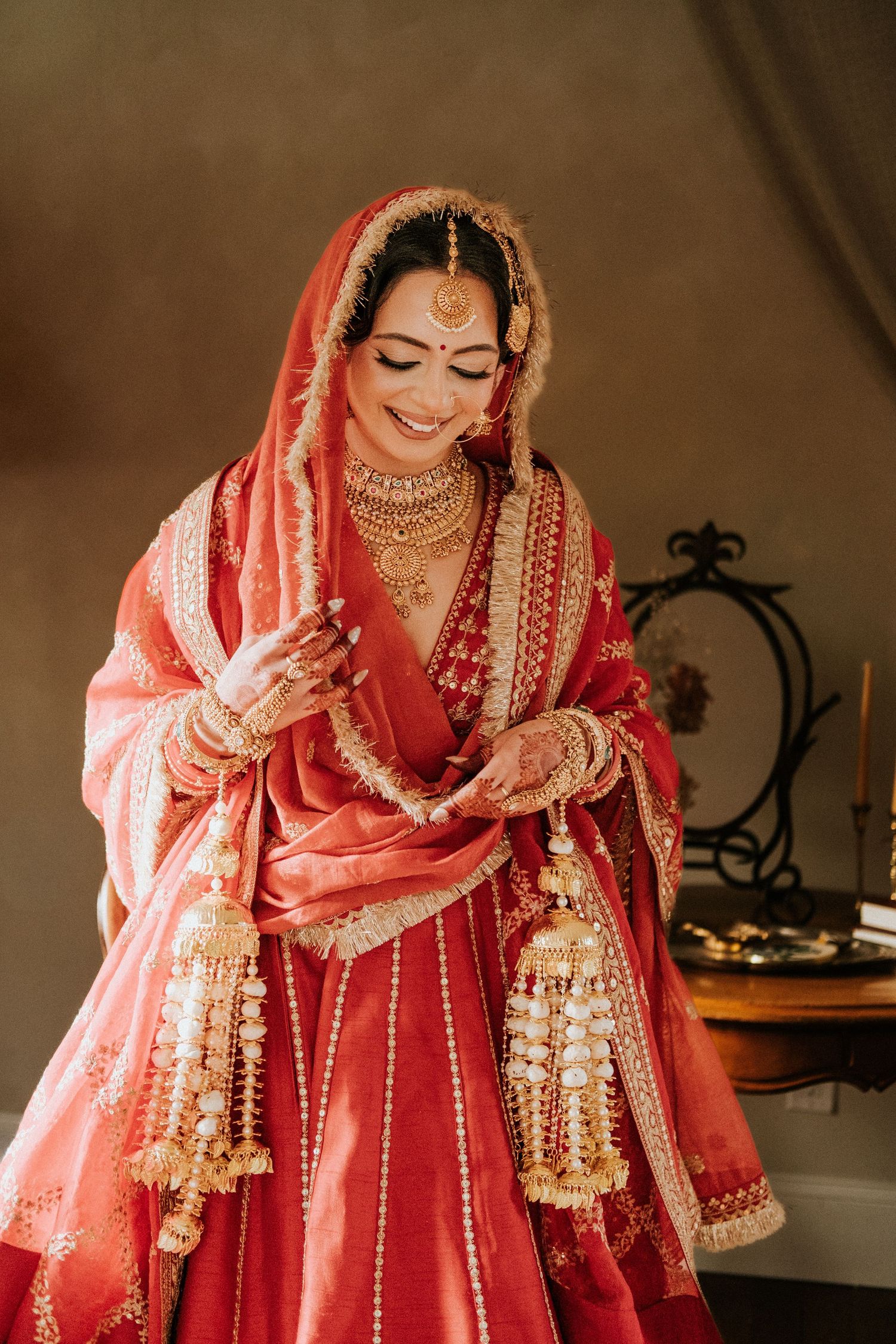 Smiling Indian bride in red lehenga with gold jewelry, maang tikka, and traditional kaleere accessories.
