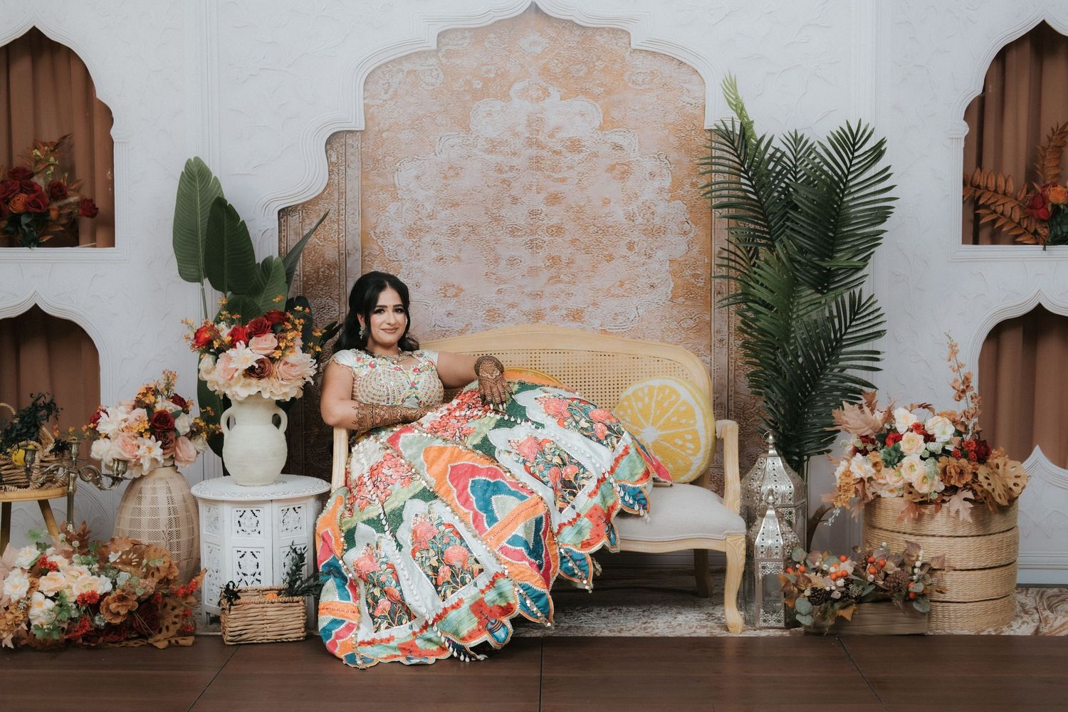 Indian bride in vibrant floral lehenga seated on ornate bench surrounded by tropical plants and flowers.