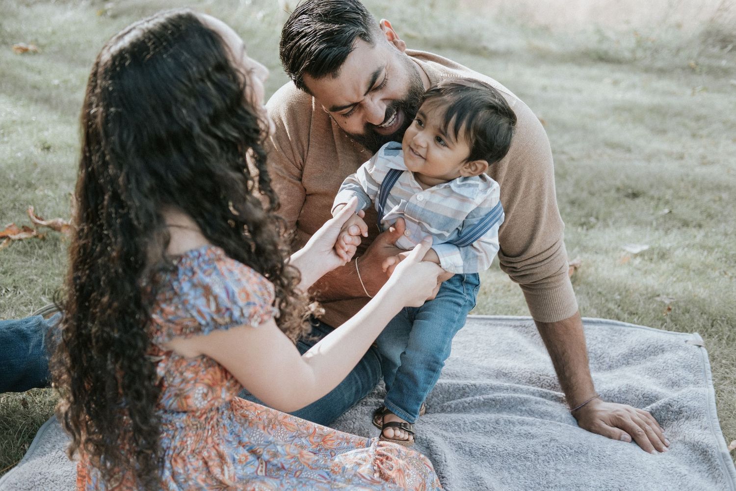 Young family sitting on blanket outdoors, parents smiling at happy baby boy in nature.