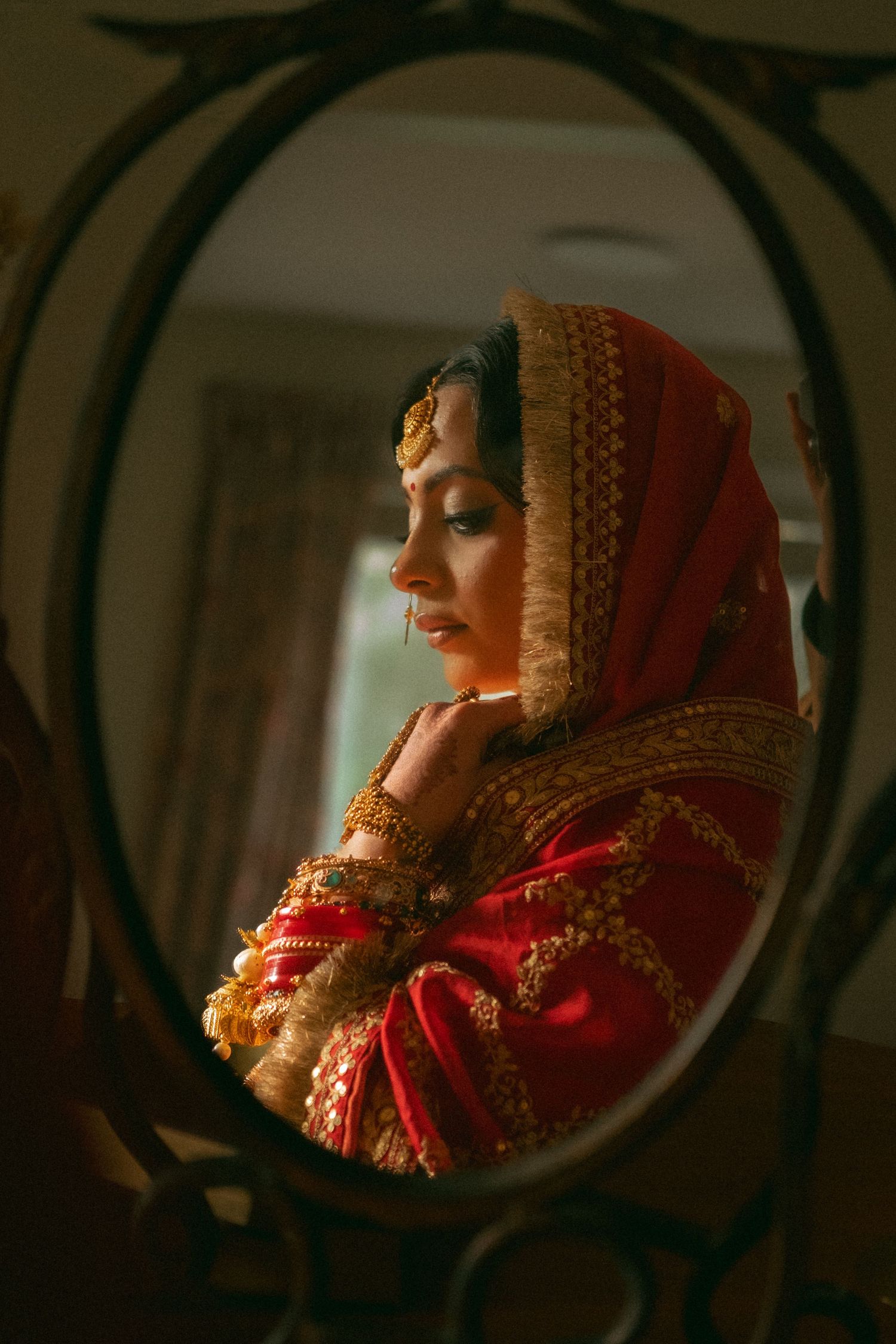 A bride in red and gold traditional South Asian bridal attire reflected in an ornate oval mirror.