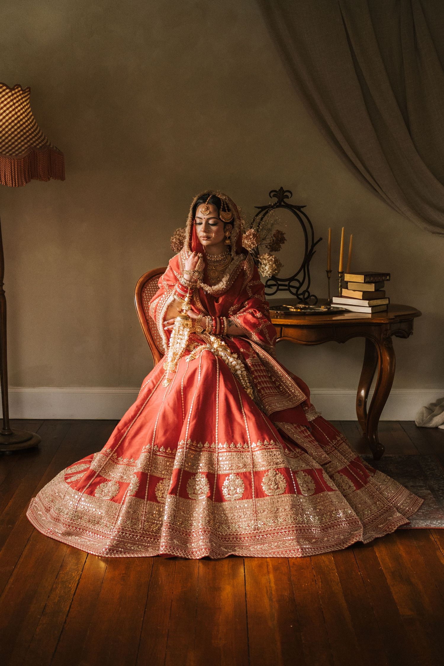 Bride in red and gold lehenga with jewelry sits gracefully in ornate chair during traditional Indian bridal photoshoot.