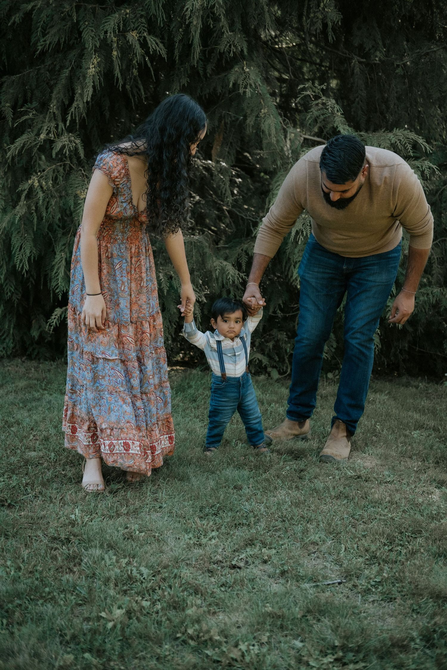 Family of three walking outdoors, parents holding toddler boy's hands in garden with evergreen trees.