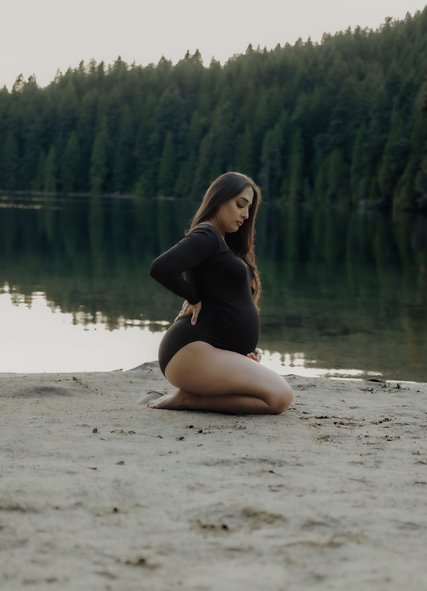 Pregnant woman in black bodysuit kneeling on sandy beach shore beside calm reflective lake.