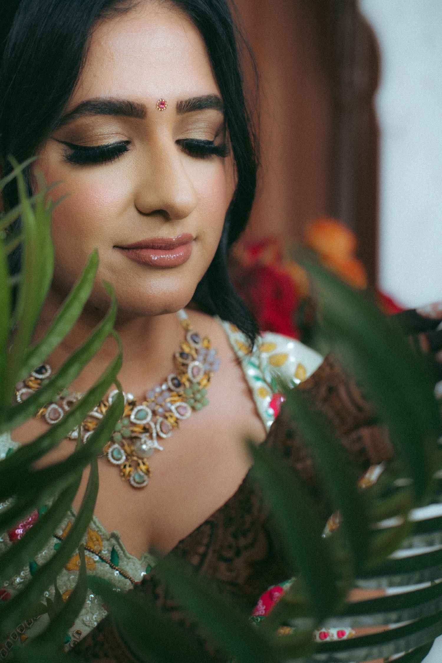 Close-up portrait of Indian bride with bindi, smoky eye makeup, and floral necklace framed by green leaves.