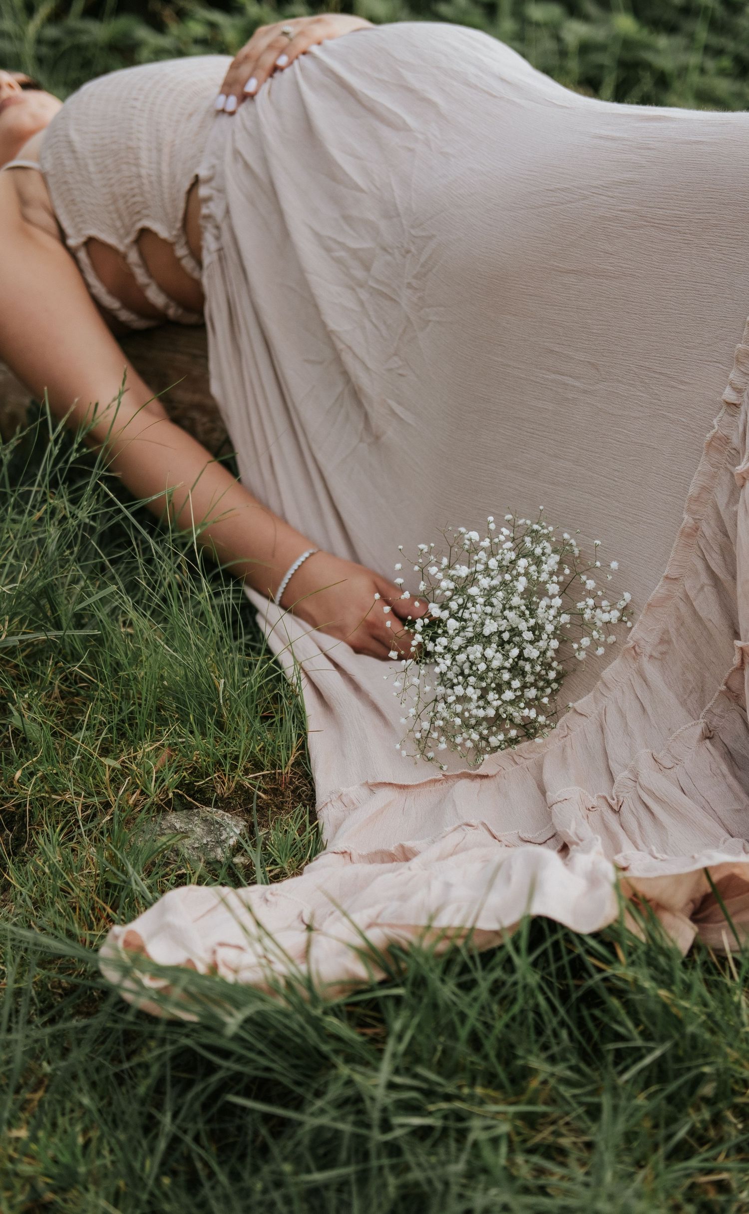 Pregnant woman in a flowing blush dress lying on grass, holding a delicate baby's breath bouquet.