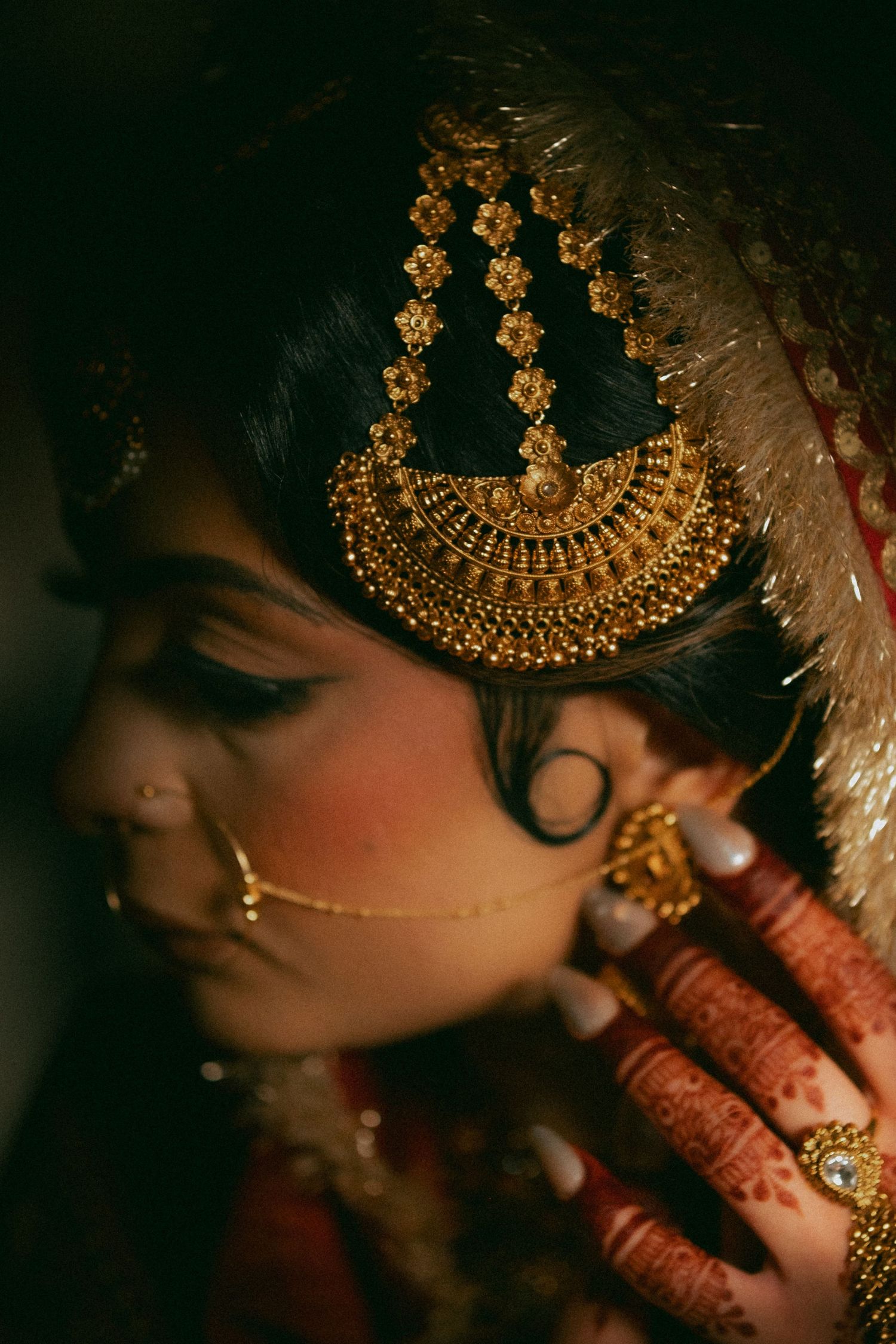 Close-up of Indian bride wearing ornate gold maang tikka jewelry and mehndi henna hands in warm light.