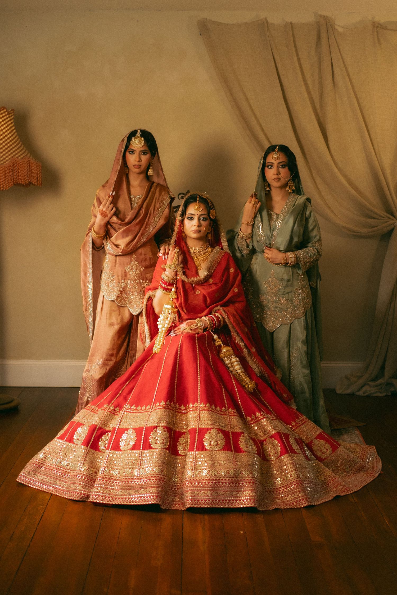 A bride in red lehenga sits flanked by two bridesmaids in traditional South Asian attire in an elegant indoor setting.