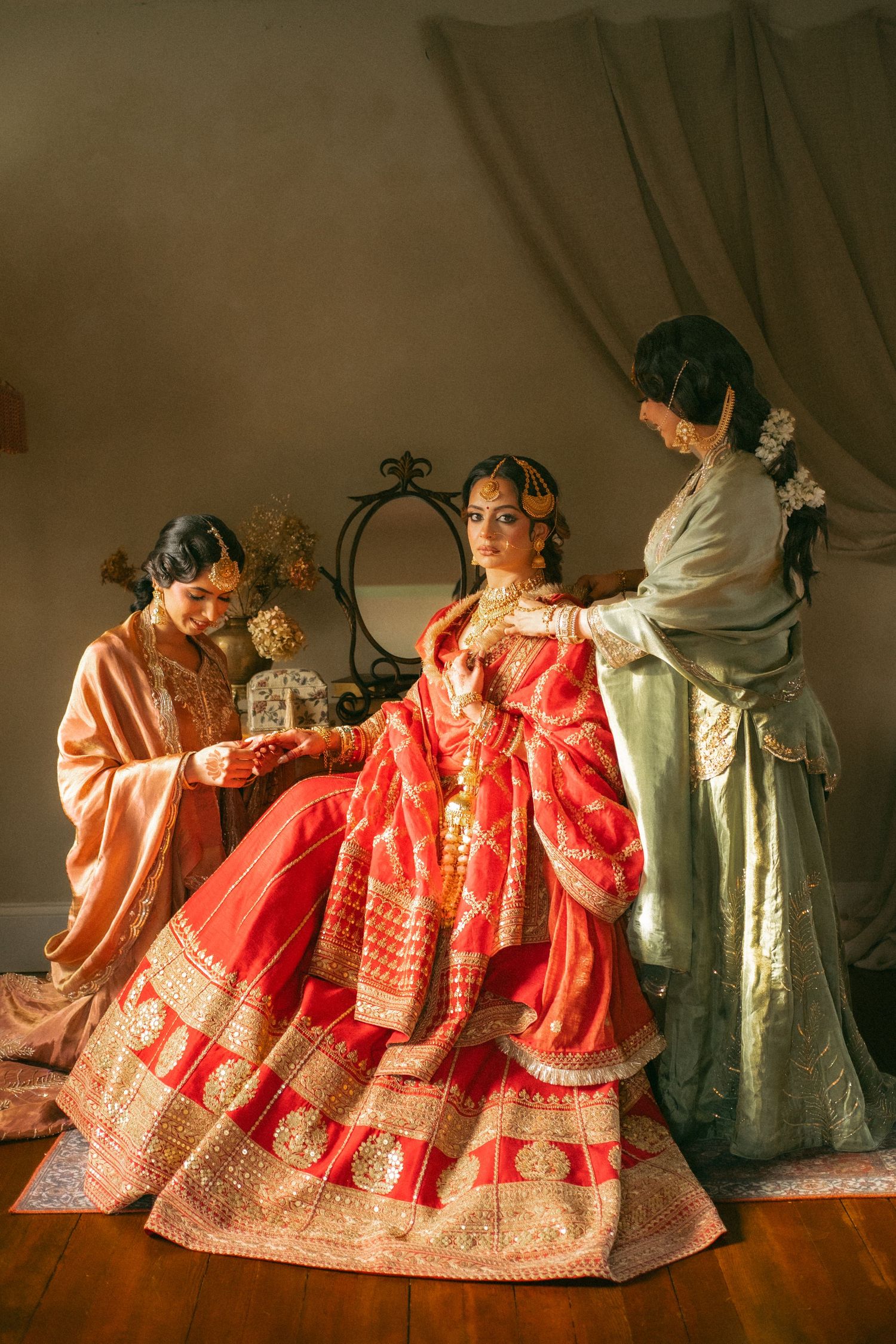 Indian bride in red lehenga being dressed by two women in traditional attire in golden light.