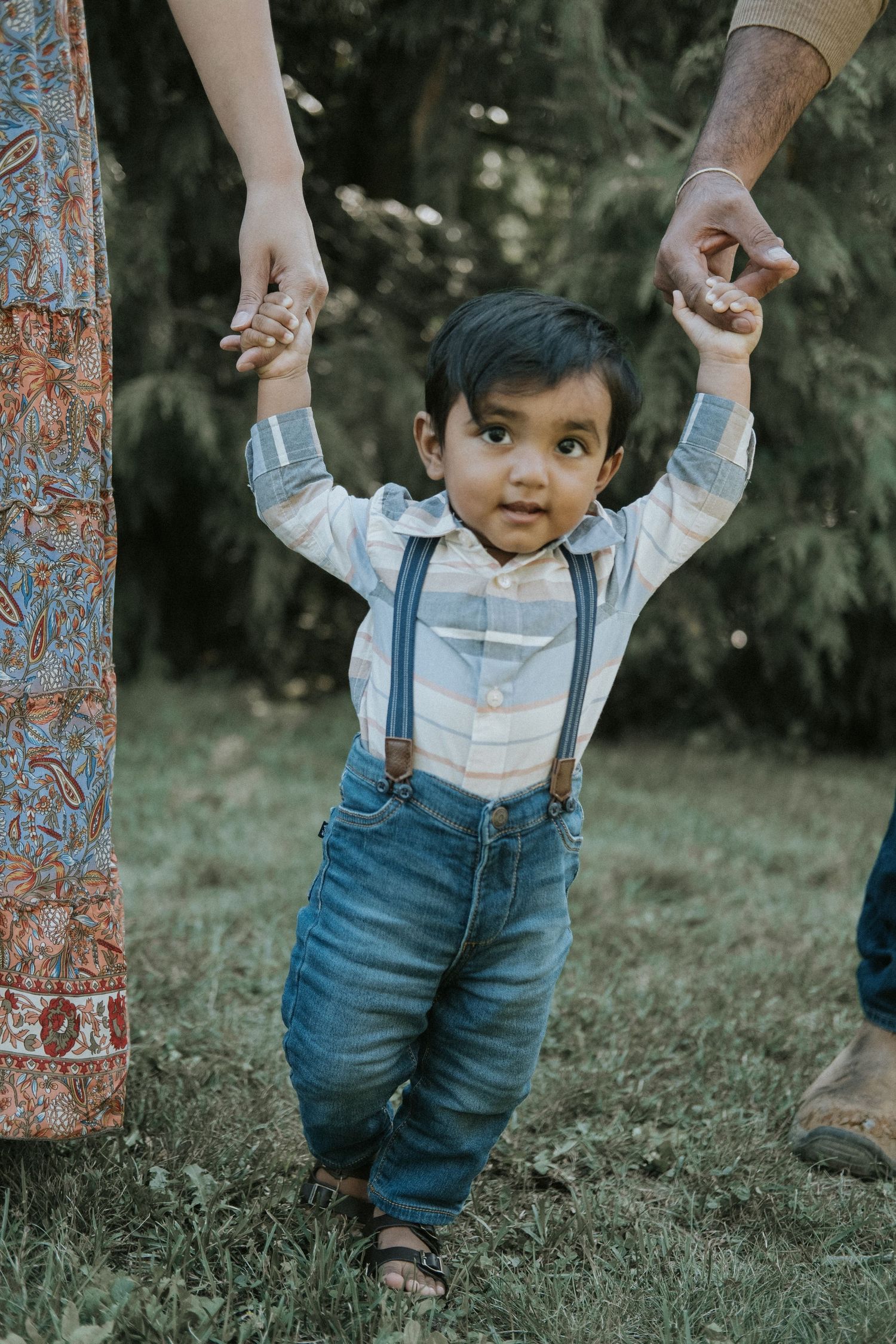 Adorable toddler boy in suspenders and jeans learning to walk with parents' helping hands outdoors.