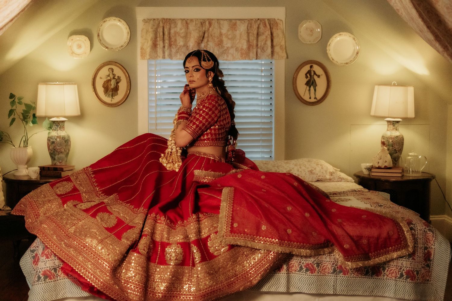 Indian bride in stunning red lehenga with gold embroidery posing on bed in warmly lit vintage bedroom.