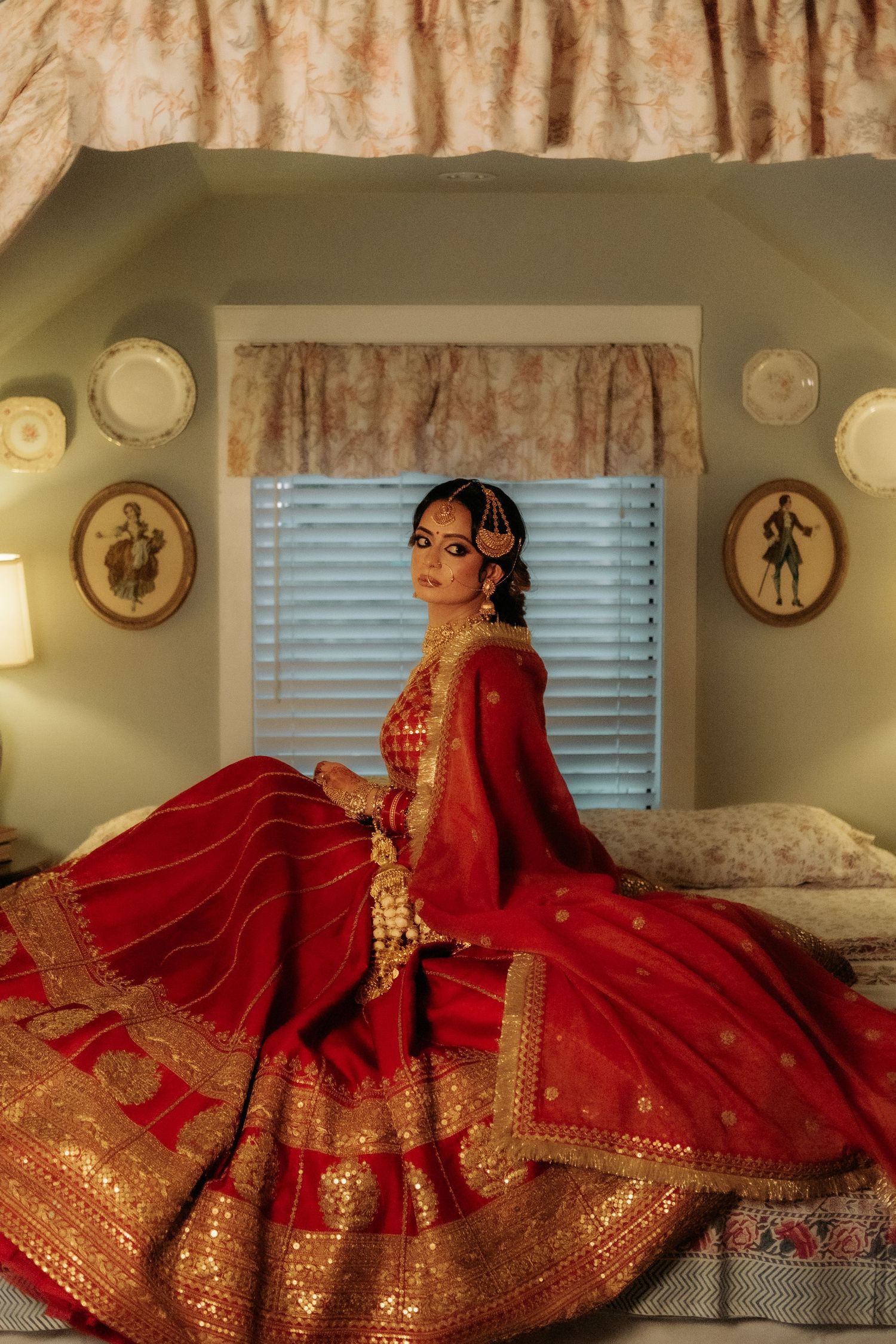 Bride in red and gold Indian bridal lehenga sitting on bed with elegant decor in background.