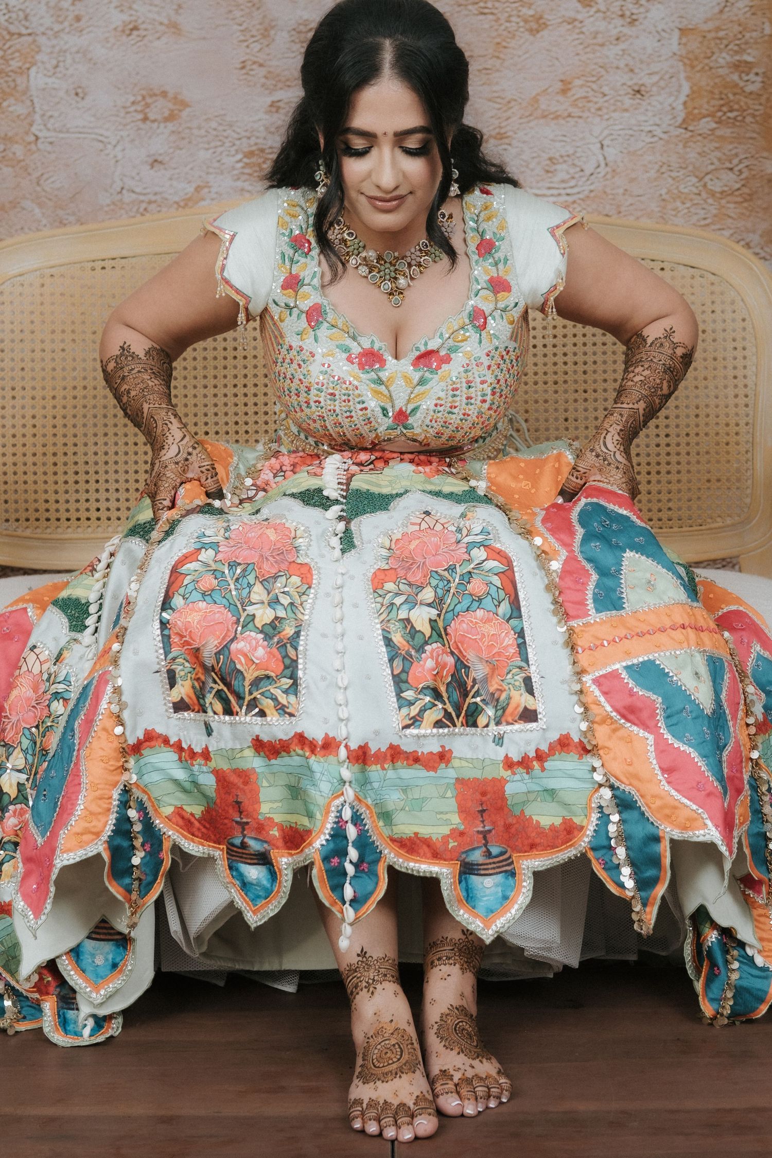 Indian bride in embroidered white floral lehenga with detailed mehndi on hands and feet seated on chair.