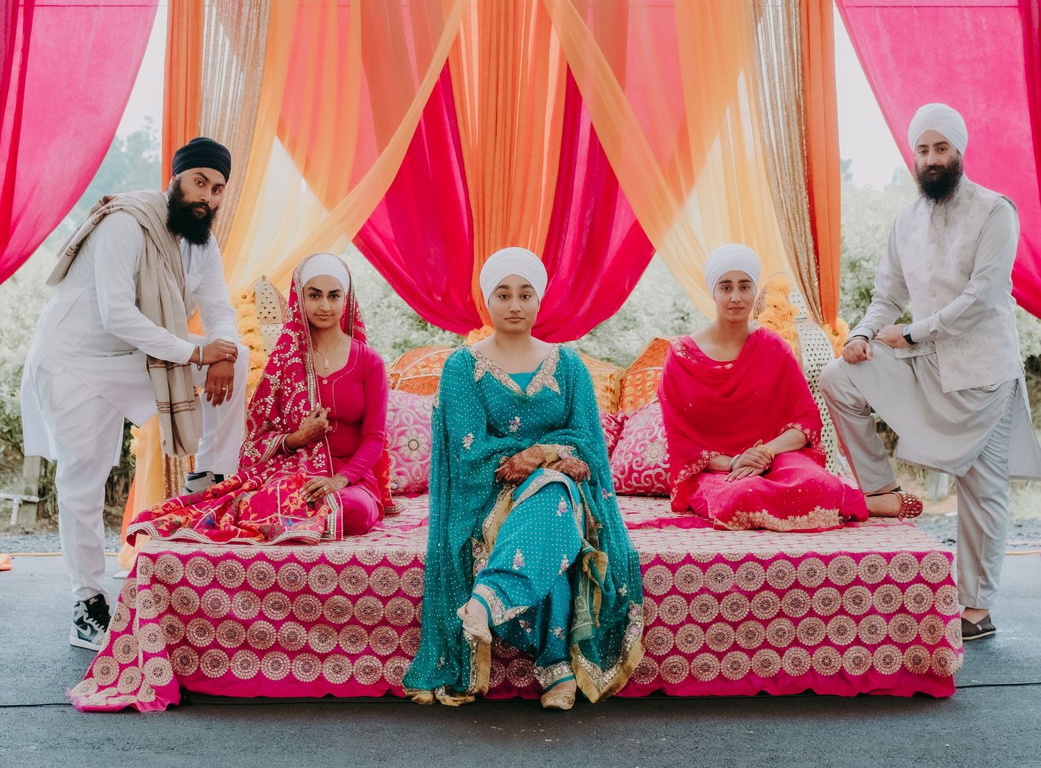 Sikh family in vibrant pink and teal traditional attire poses at colorful outdoor wedding ceremony.