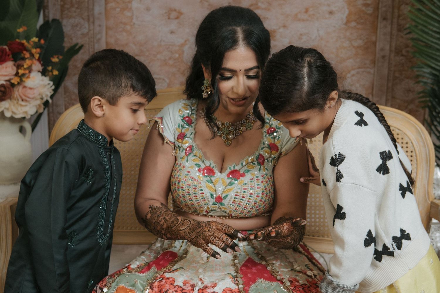 Indian bride in floral lehenga with mehndi hands seated between two children at wedding celebration.