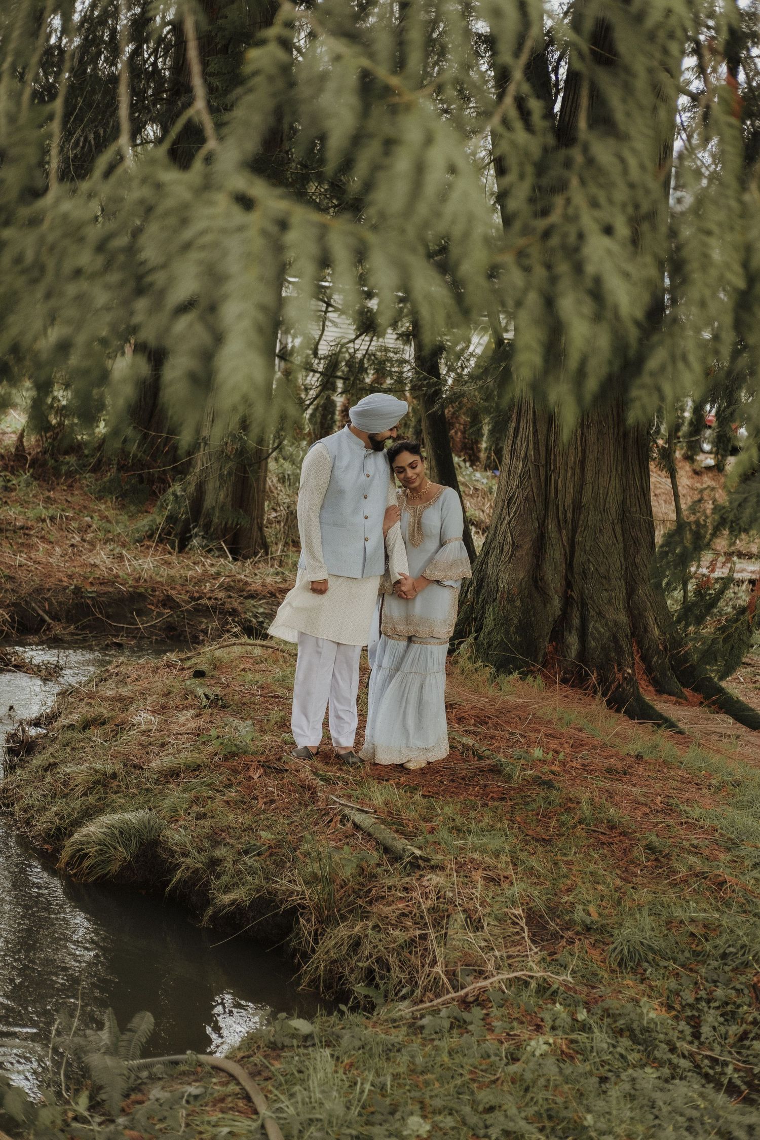Sikh couple in white traditional attire standing near a stream in a lush forest setting.