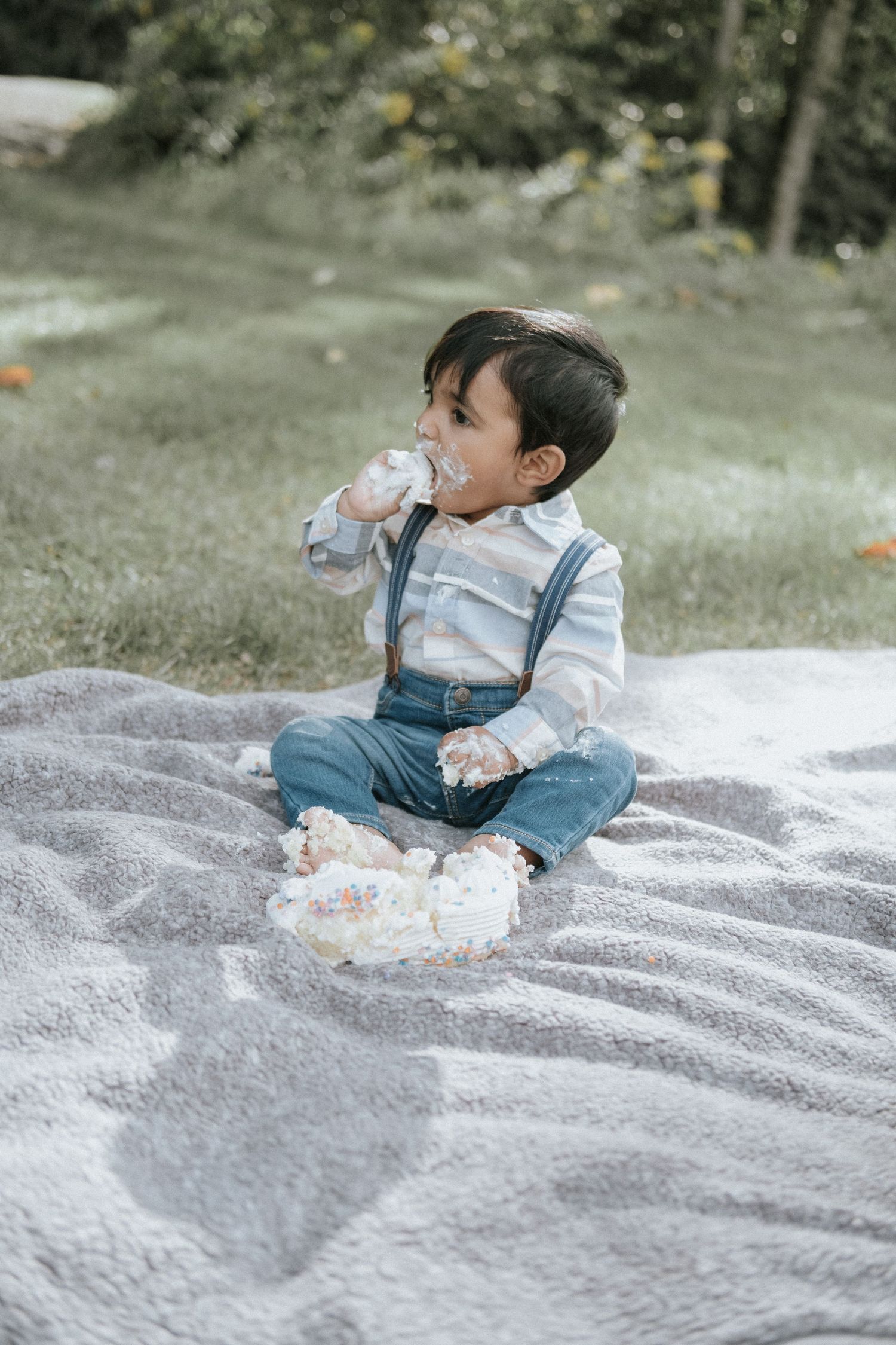 Baby boy in suspenders sitting on blanket outdoors enjoying messy first birthday cake smash.