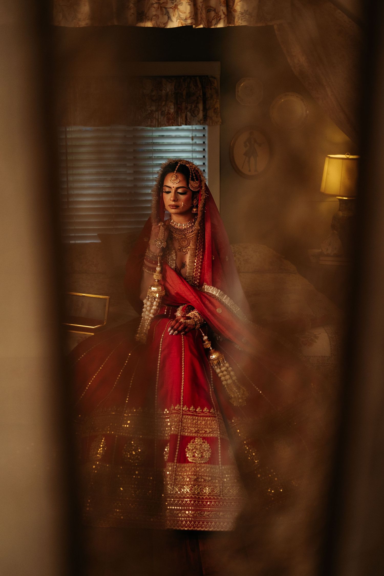 Indian bride in red and gold lehenga standing elegantly in warmly lit room framed by curtains.