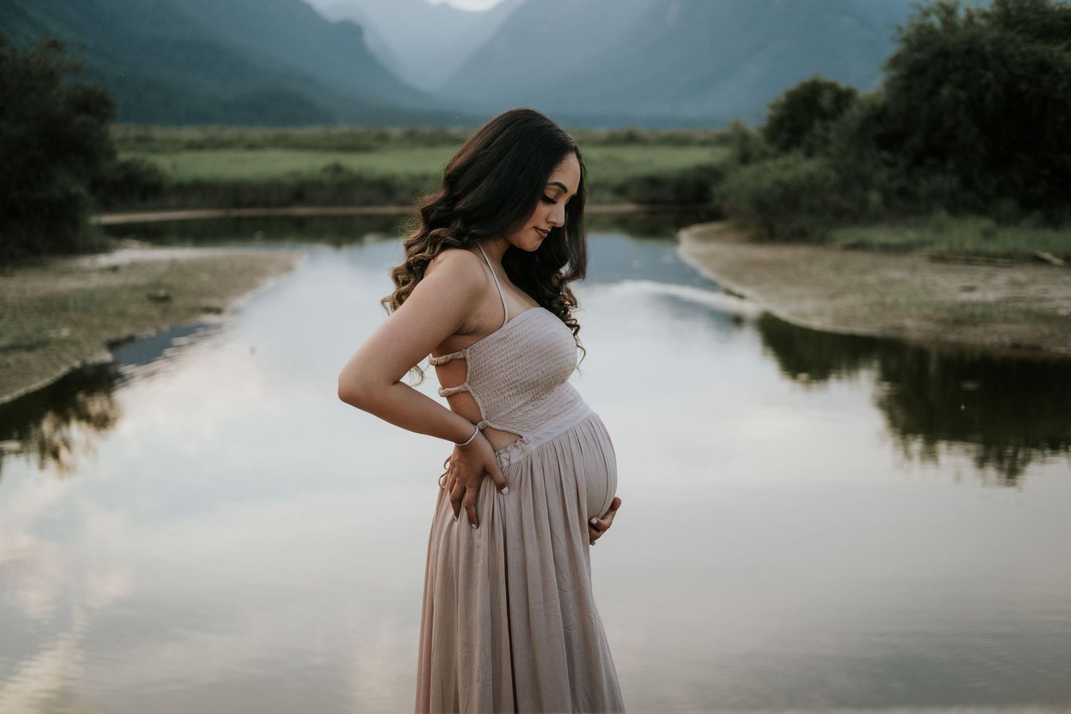 Pregnant woman in elegant blush dress cradling her baby bump beside a tranquil mountain lake.