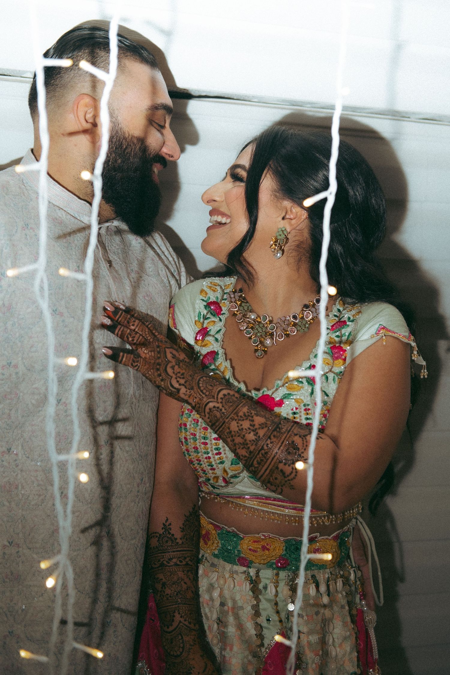 Indian couple sharing a romantic moment amid string lights, bride in embroidered lehenga with mehndi-adorned hands.