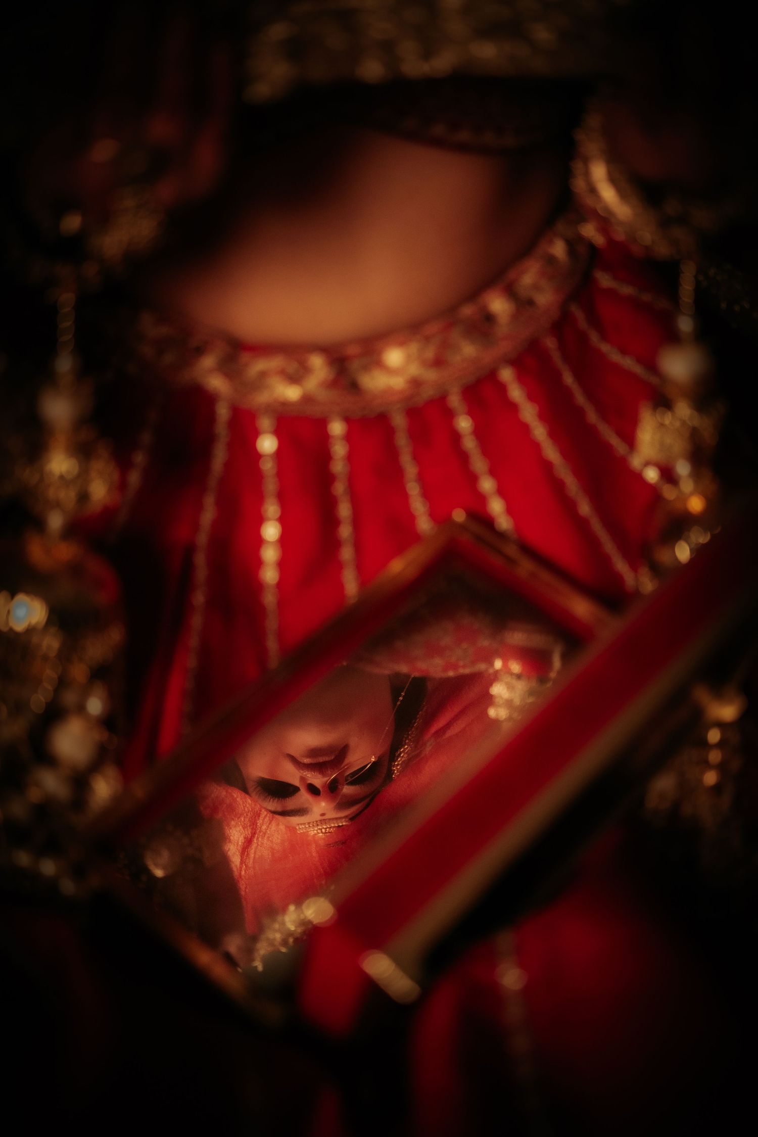 Close-up of a bride in red and gold bridal attire holding a decorative box with wedding rings in dim light.