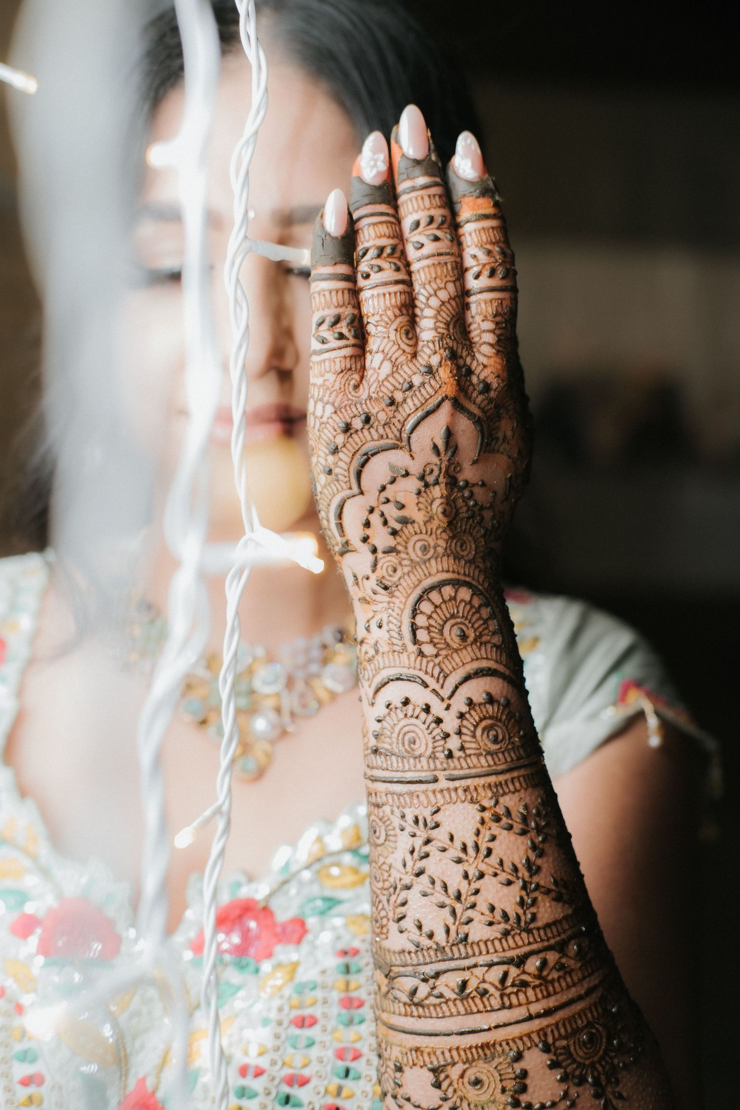 Indian bride with intricate mehndi henna on hand posing behind string lights in colorful bridal outfit.