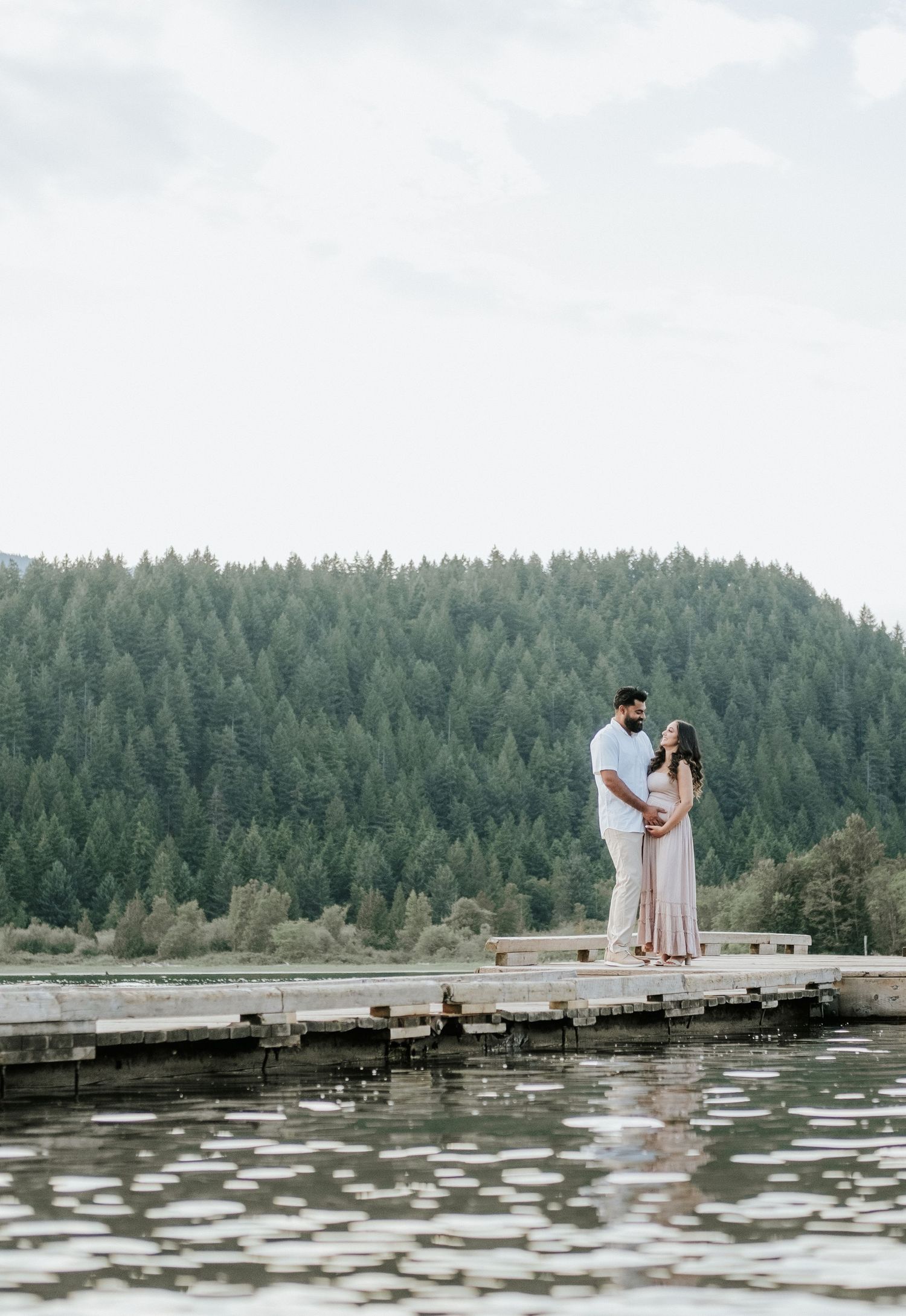 Expecting couple standing on a wooden dock, embracing against a backdrop of forested mountains and calm water.
