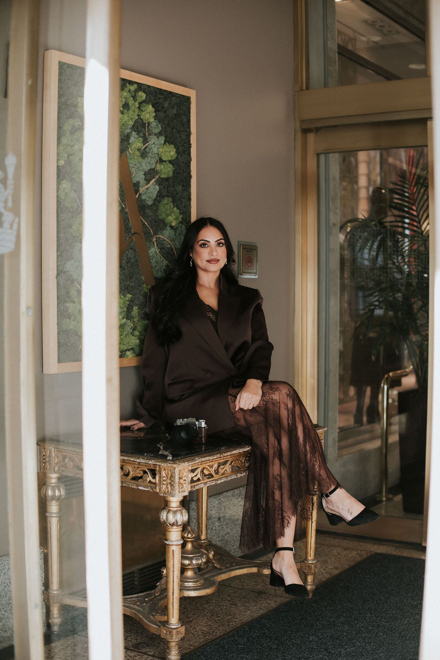Elegant brunette woman in brown blazer seated on ornate gold table in upscale lobby.