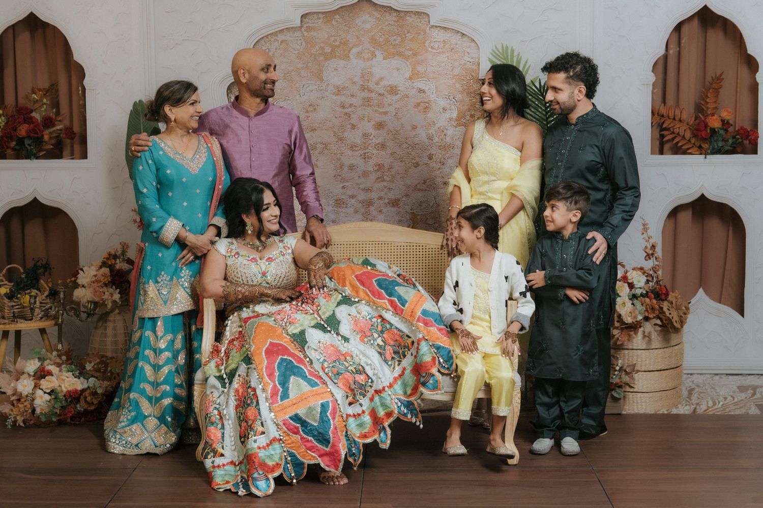 A South Asian family in vibrant traditional outfits poses together around a bride at a wedding event.