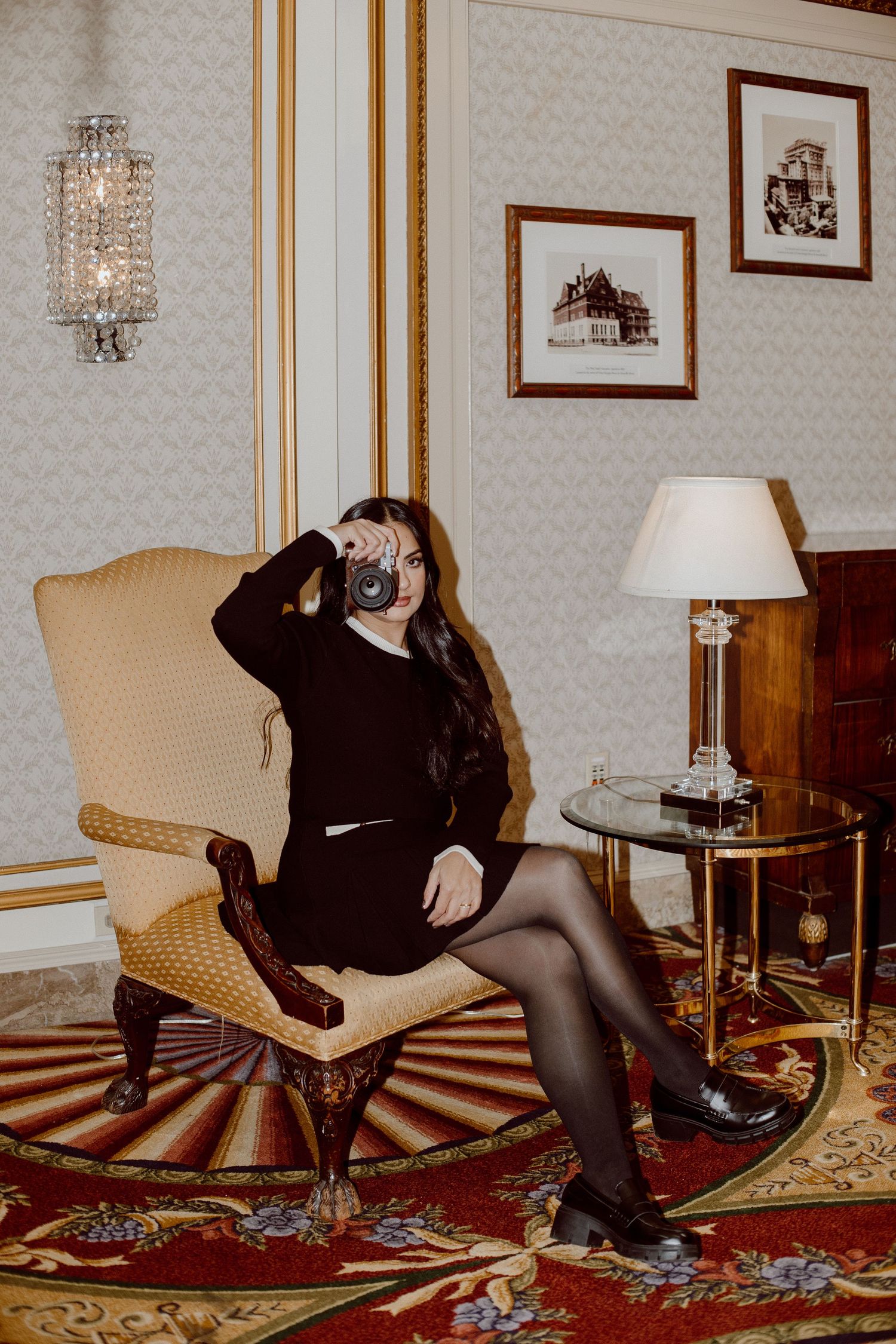 Woman in black outfit sitting in elegant chair photographing with vintage camera indoors.