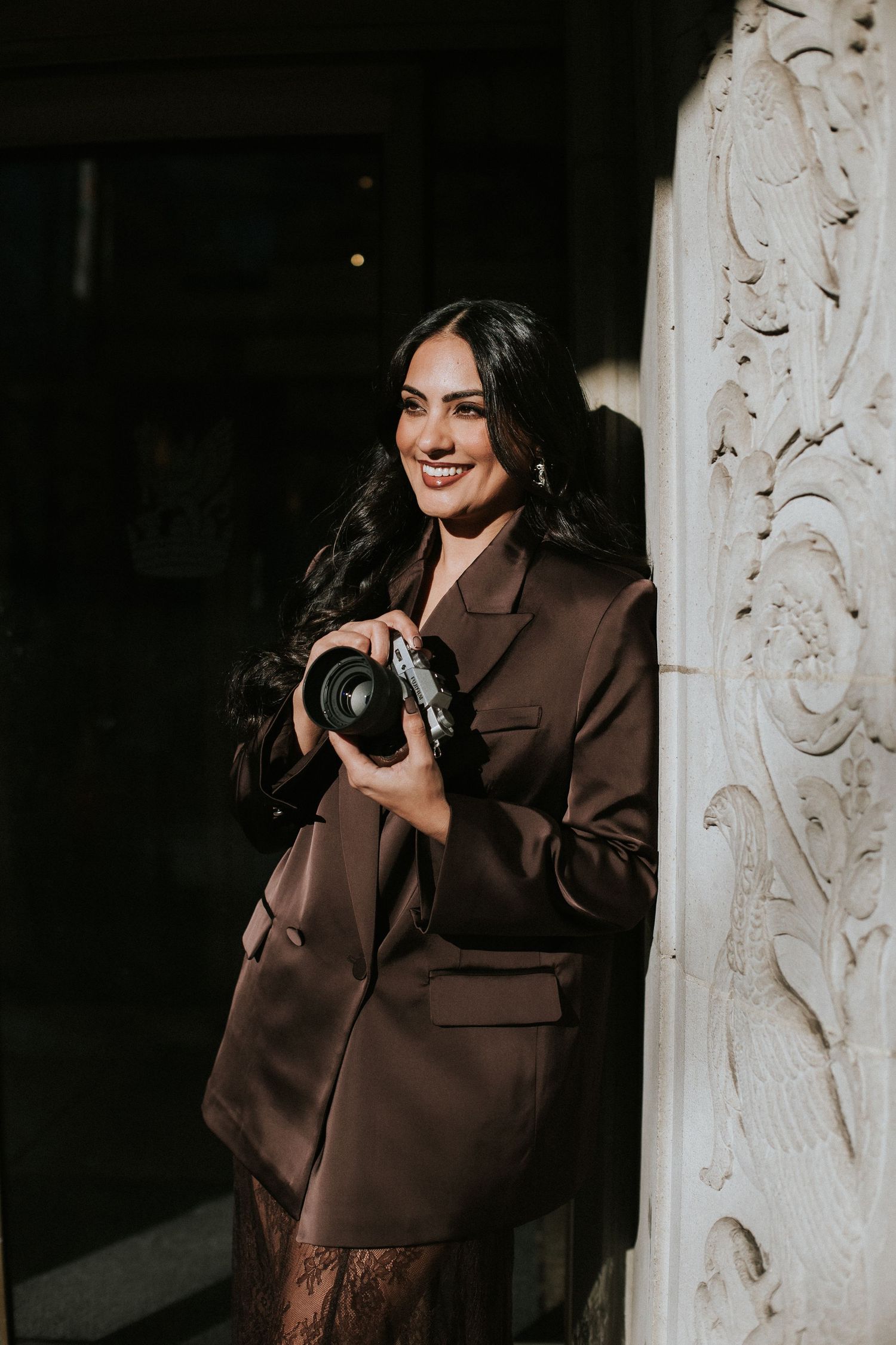 Smiling woman in brown blazer holding a camera outdoors near ornate white column.