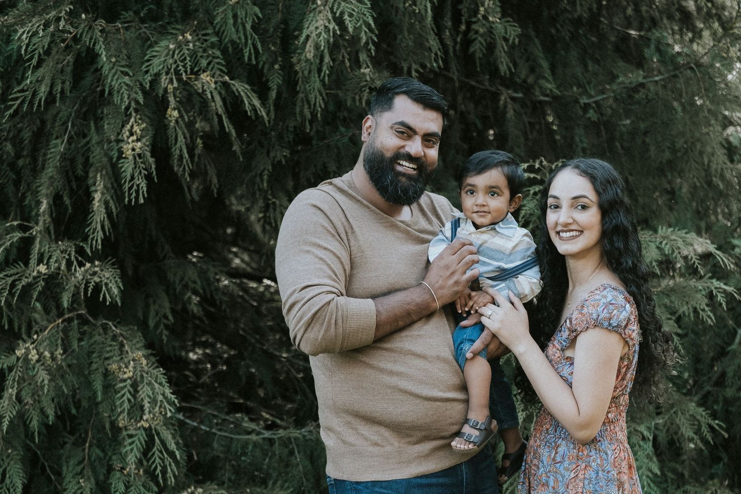 Happy South Asian family portrait with bearded father holding baby boy and smiling mother outdoors.