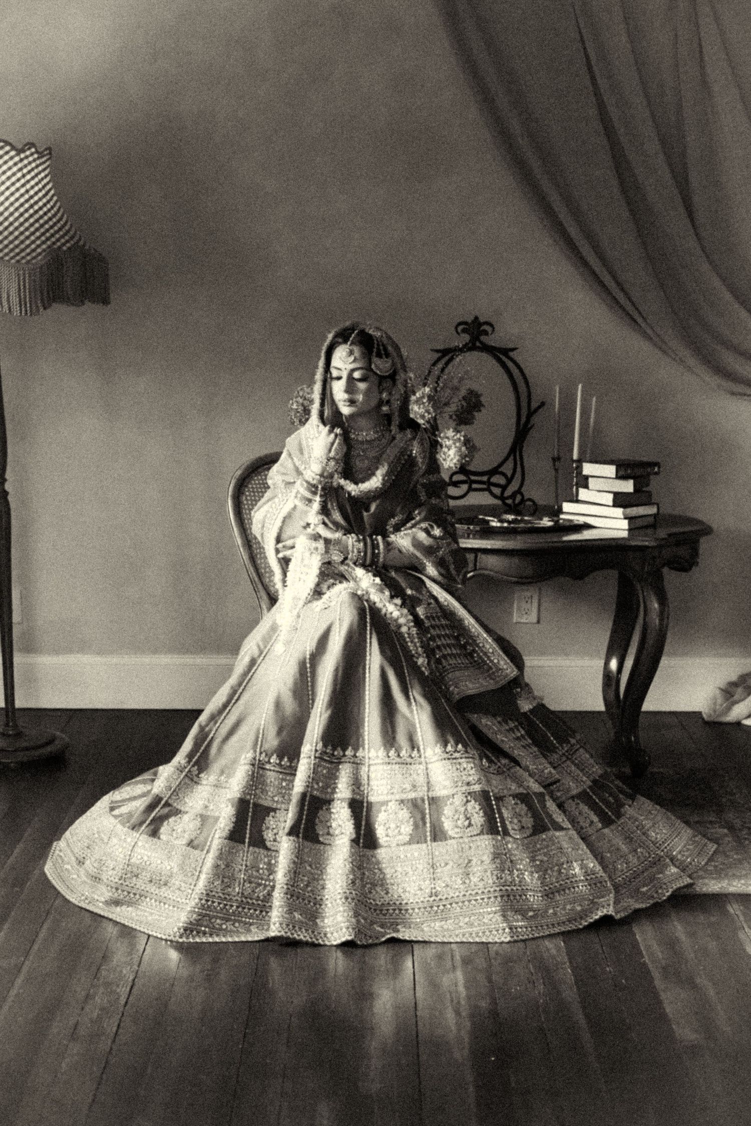 Vintage sepia portrait of Indian bride in embroidered lehenga seated elegantly by antique desk.