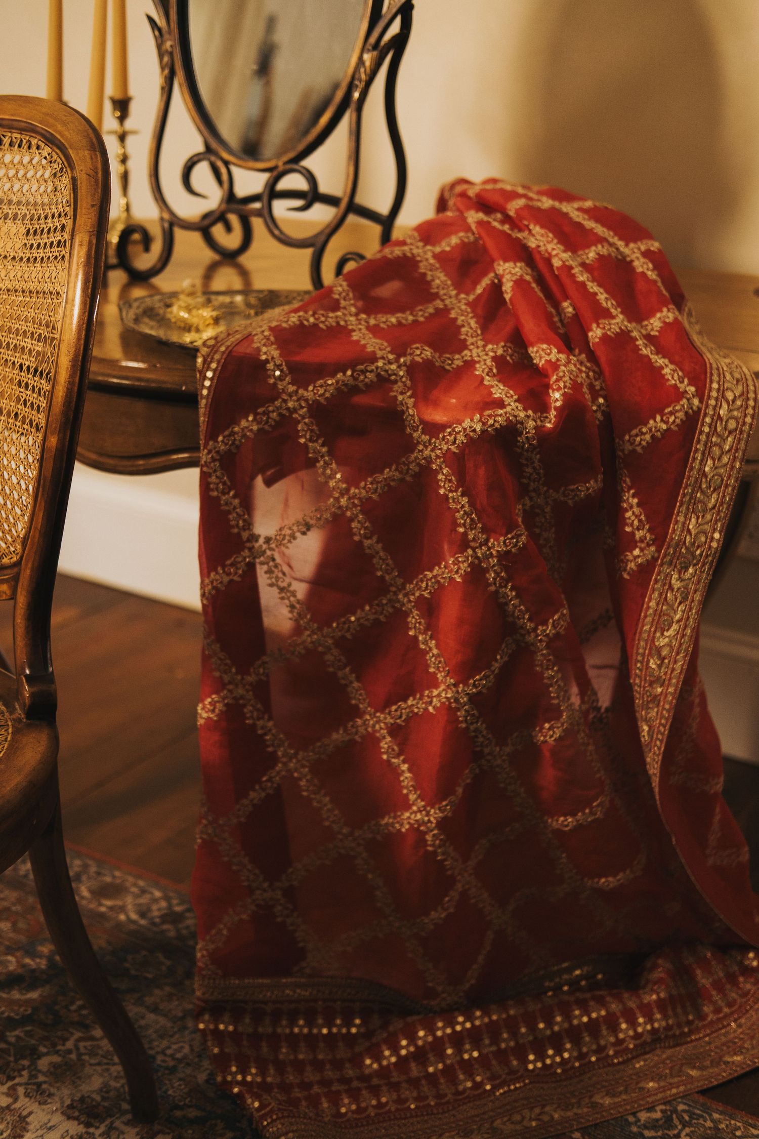 Red silk saree with gold embroidery draped over a vintage cane chair near a vanity mirror.
