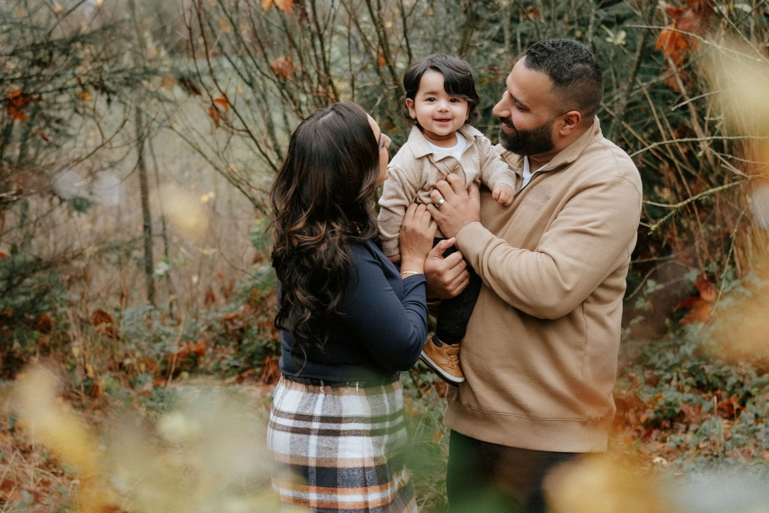 A happy family with a smiling toddler enjoys an autumn outdoor portrait surrounded by colorful fall foliage.