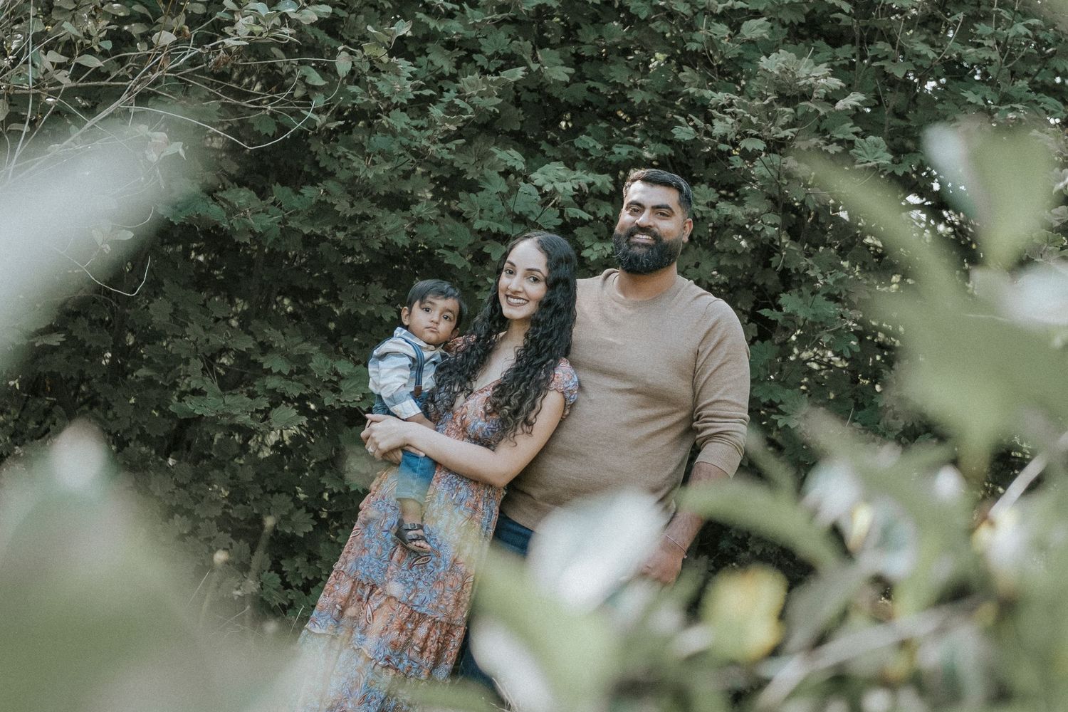 A happy South Asian family of three poses together surrounded by green foliage in a natural outdoor setting.