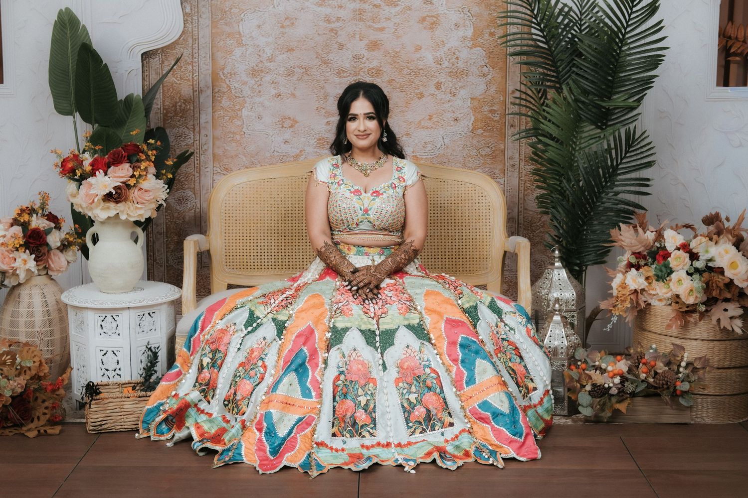 Indian bride in colorful floral lehenga seated gracefully on decorative bench with tropical wedding decor.