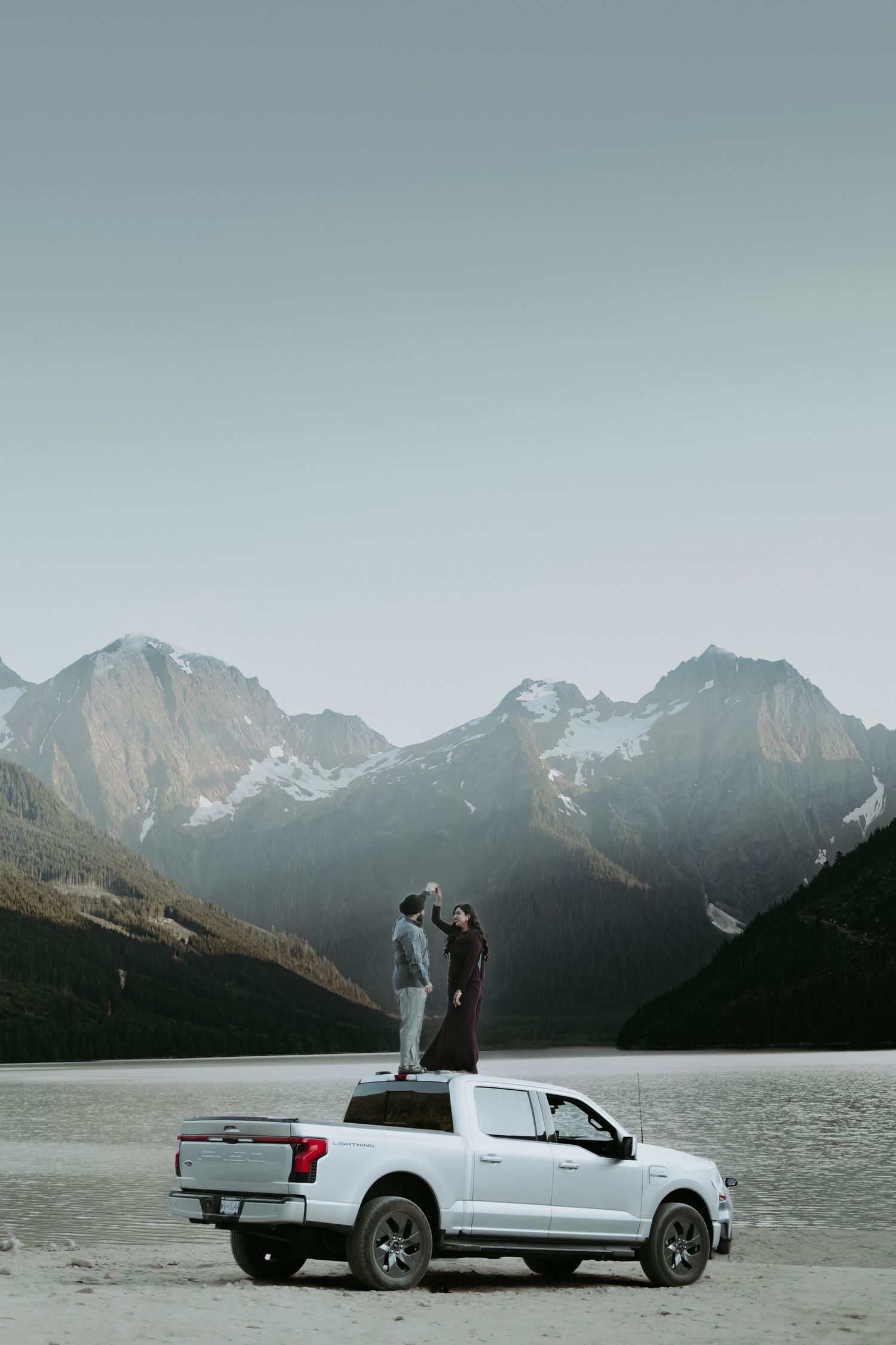 Couple standing on white Ford pickup truck bed with stunning snow-capped mountain range backdrop in wilderness.