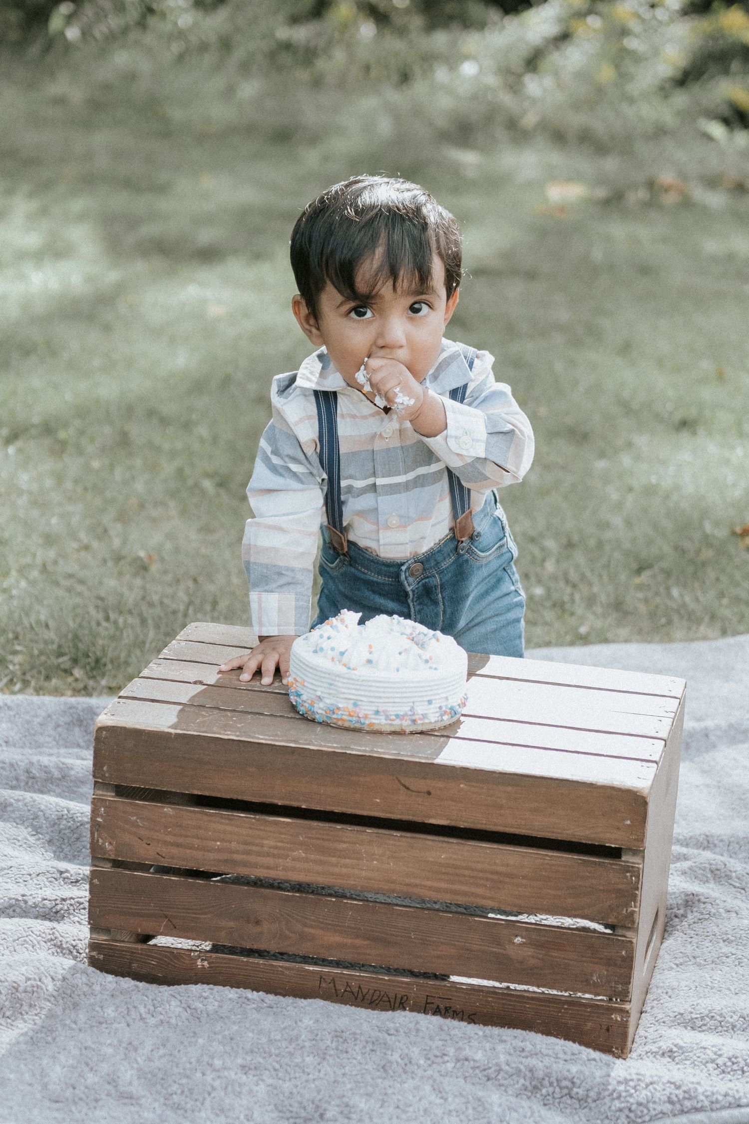 Toddler boy in suspenders standing at wooden crate eating first birthday smash cake outdoors.