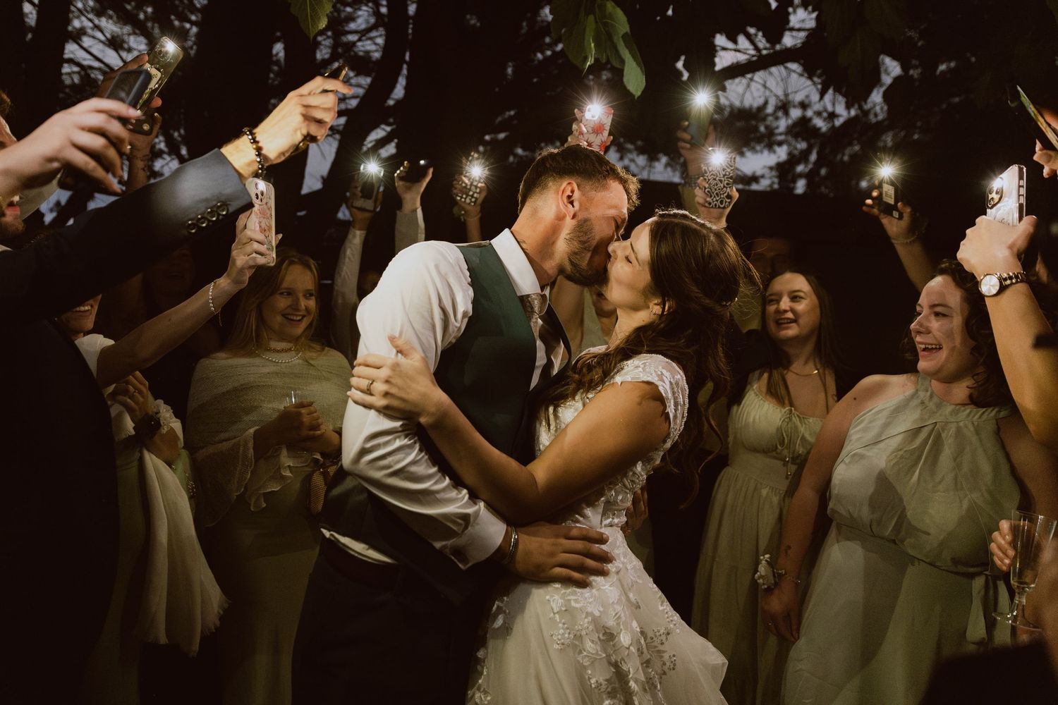 Un couple de mariés fait un trend photo de mariage avec leur amis que les éclairent avec leur téléphone pendant qu'ils s'embrassent.